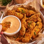A hand dips a crispy fried chicken tender into a bowl of creamy dipping sauce. The tenders are garnished with chopped herbs and served in a paper-lined bowl on a stone surface.
