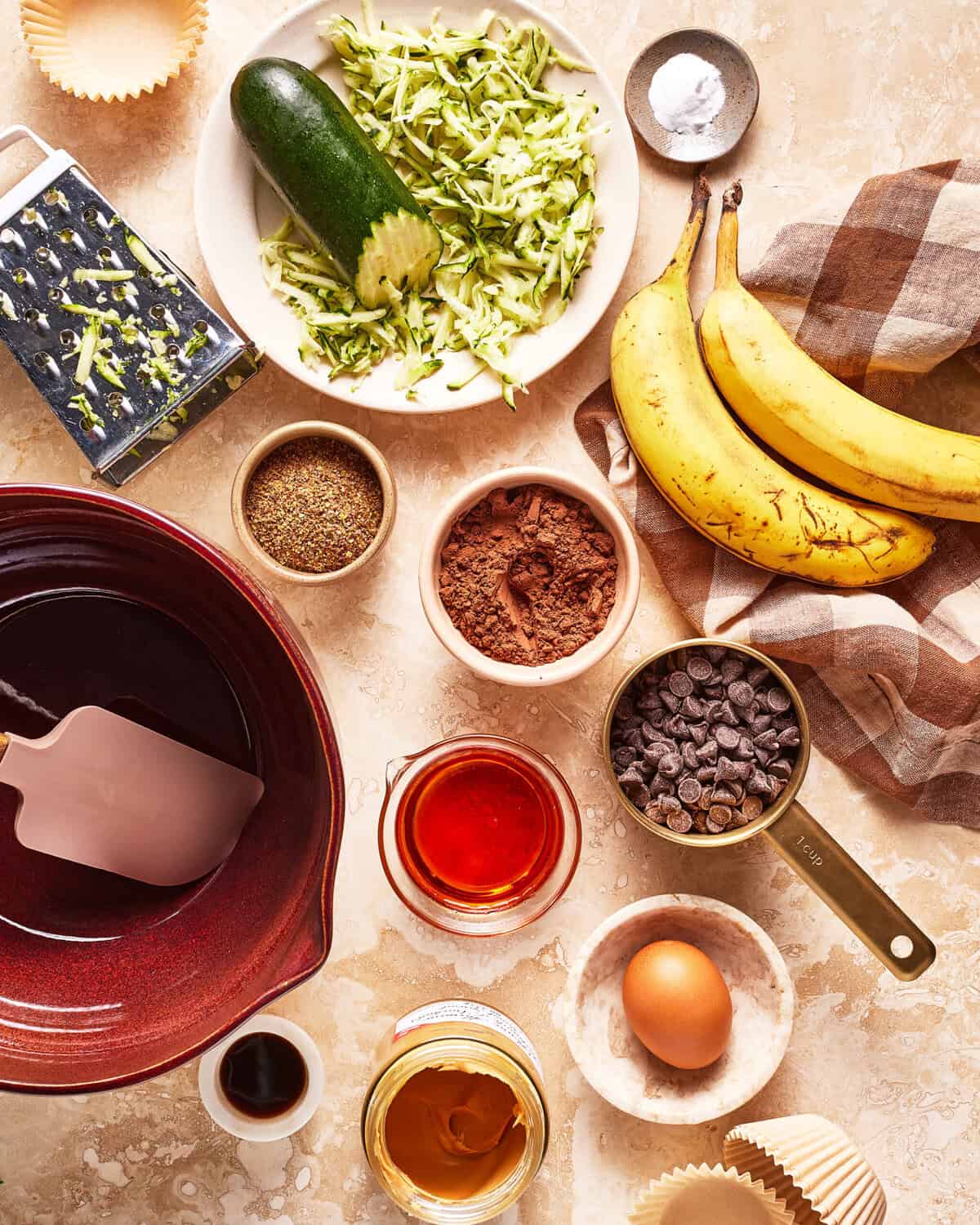 Baking ingredients on a counter, including grated zucchini, bananas, cocoa powder, chocolate chips, an egg, flaxseed, peanut butter, vanilla, oil, and a mixing bowl with a spatula.