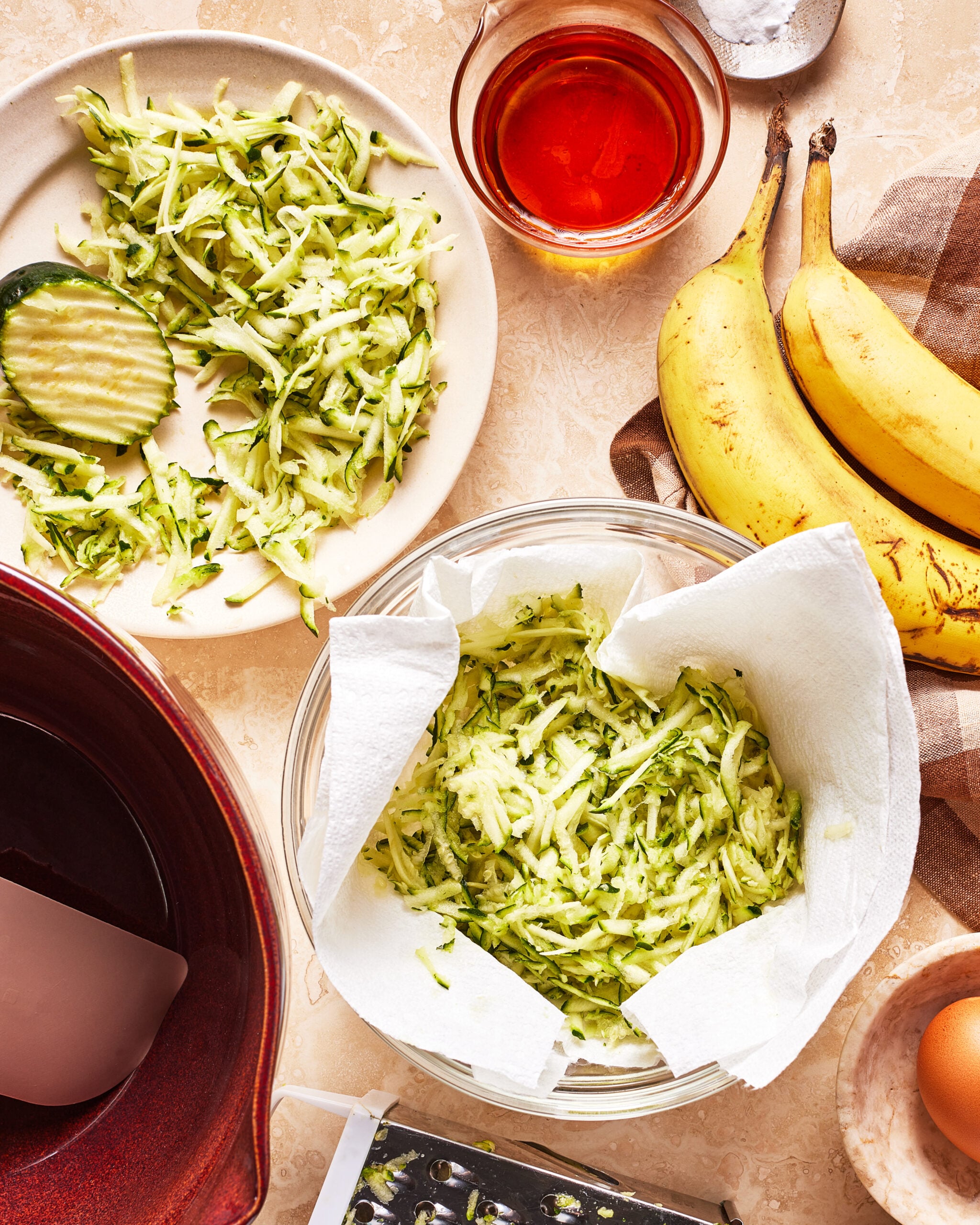 Overhead view of shredded zucchini on a plate and in a paper towel-lined bowl, bananas, an egg, grated zucchini, a glass of amber liquid, a red bowl, and a grater on a light countertop.