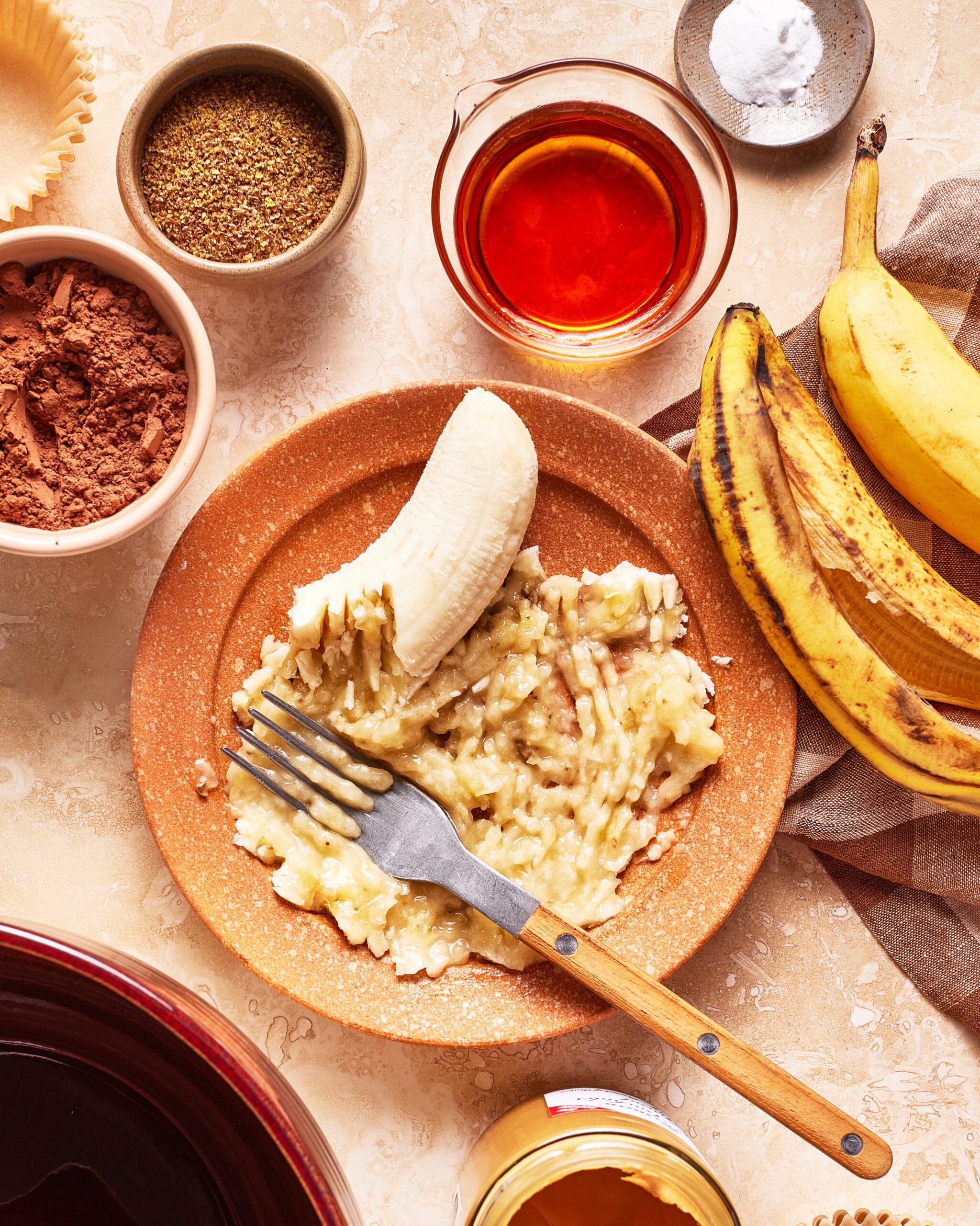 A fork mashing a banana on a plate surrounded by whole bananas, cocoa powder, flaxseed, baking soda, syrup, and a jar of honey on a beige surface.