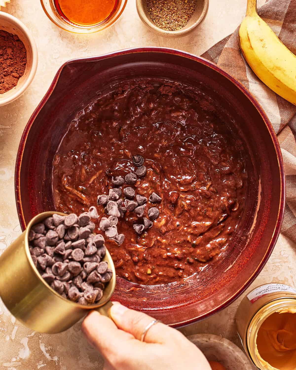 A hand pours chocolate chips into a bowl of chocolate batter. Surrounding the bowl are ingredients like a banana, cocoa powder, and honey on a kitchen countertop.