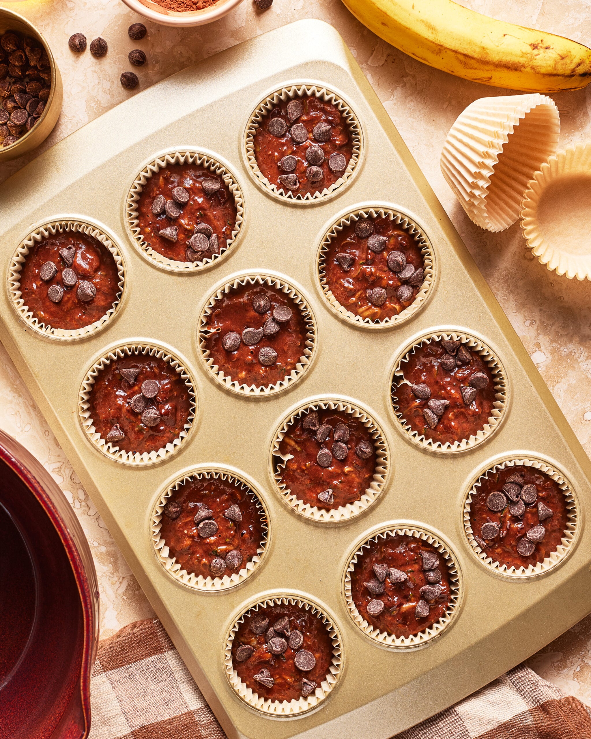 A muffin tin with 12 unbaked chocolate chip banana muffins in paper liners. Nearby are chocolate chips, a peeled banana, empty liners, a red bowl, and a checkered cloth.
