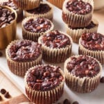 Chocolate muffins with chocolate chips arranged on a white surface, some stacked, with a cup of chocolate chips nearby. Muffins have crinkled paper liners and a rich, moist appearance.