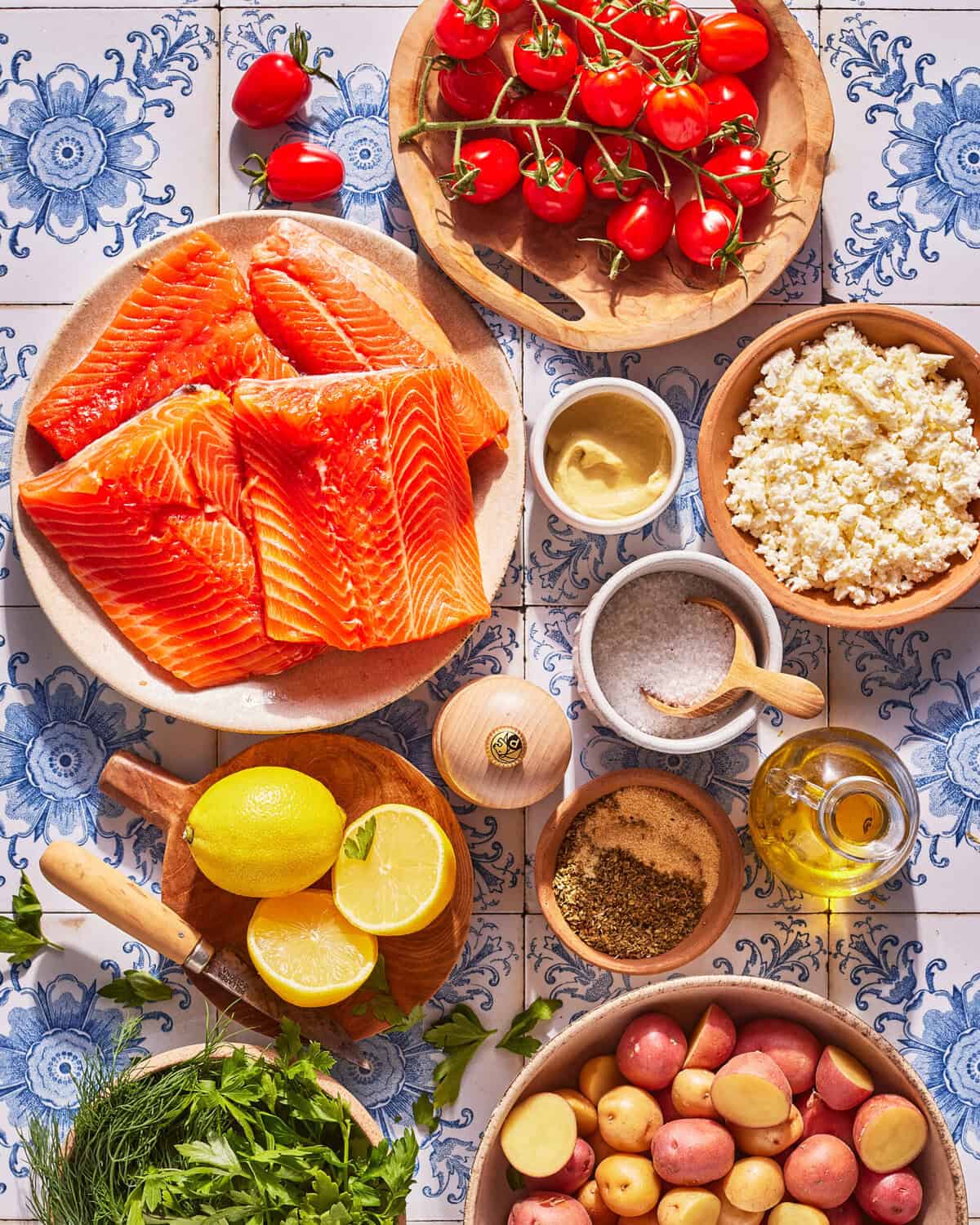 Fresh salmon fillets, cherry tomatoes, cauliflower rice, red potatoes, lemon, parsley, olive oil, mustard, salt, pepper, and an onion arranged on blue and white patterned tile.