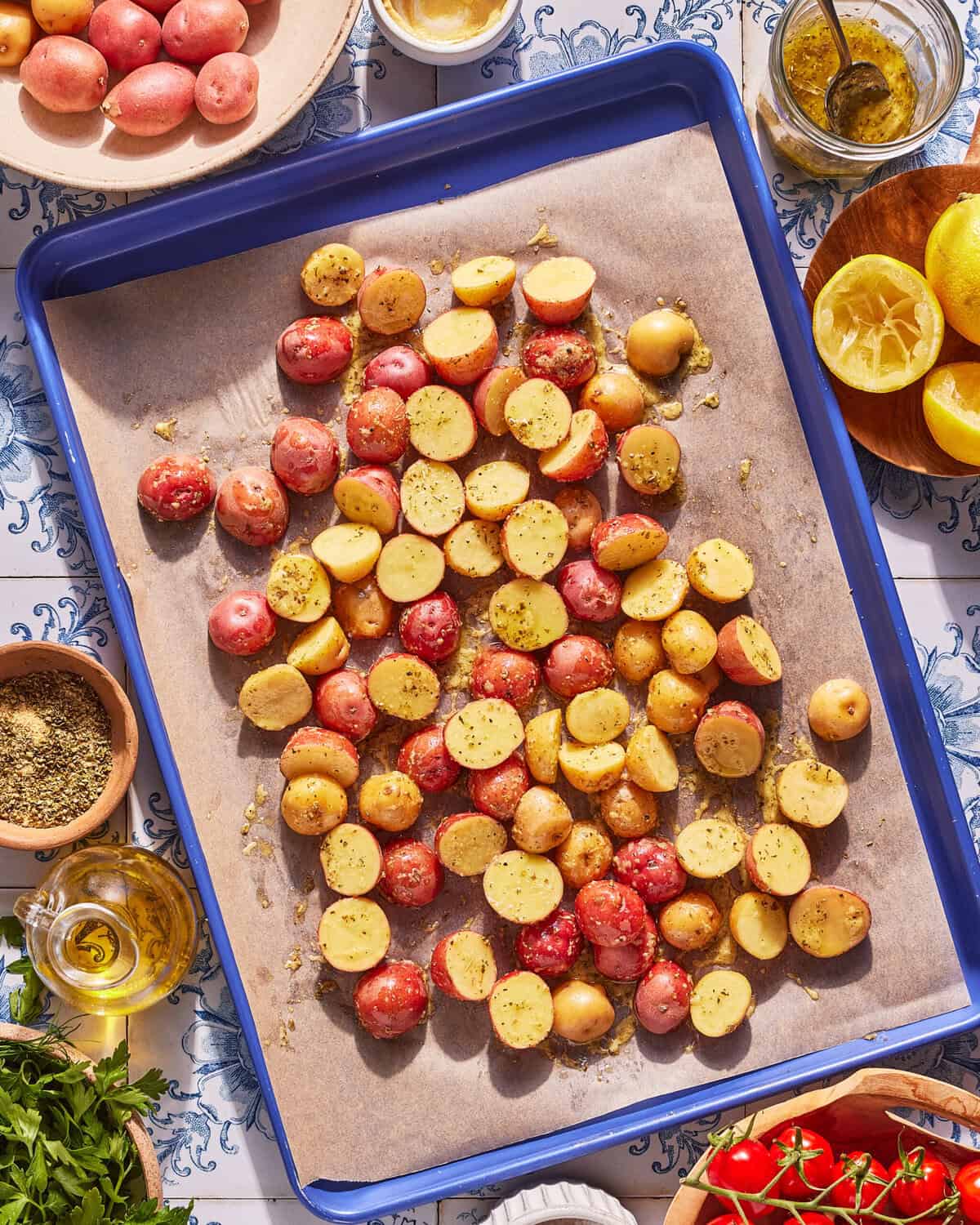 A baking tray with halved baby red and yellow potatoes, sprinkled with herbs, sits on parchment paper. Surrounding the tray are bowls of oil, spices, lemons, and fresh tomatoes on a patterned tablecloth.