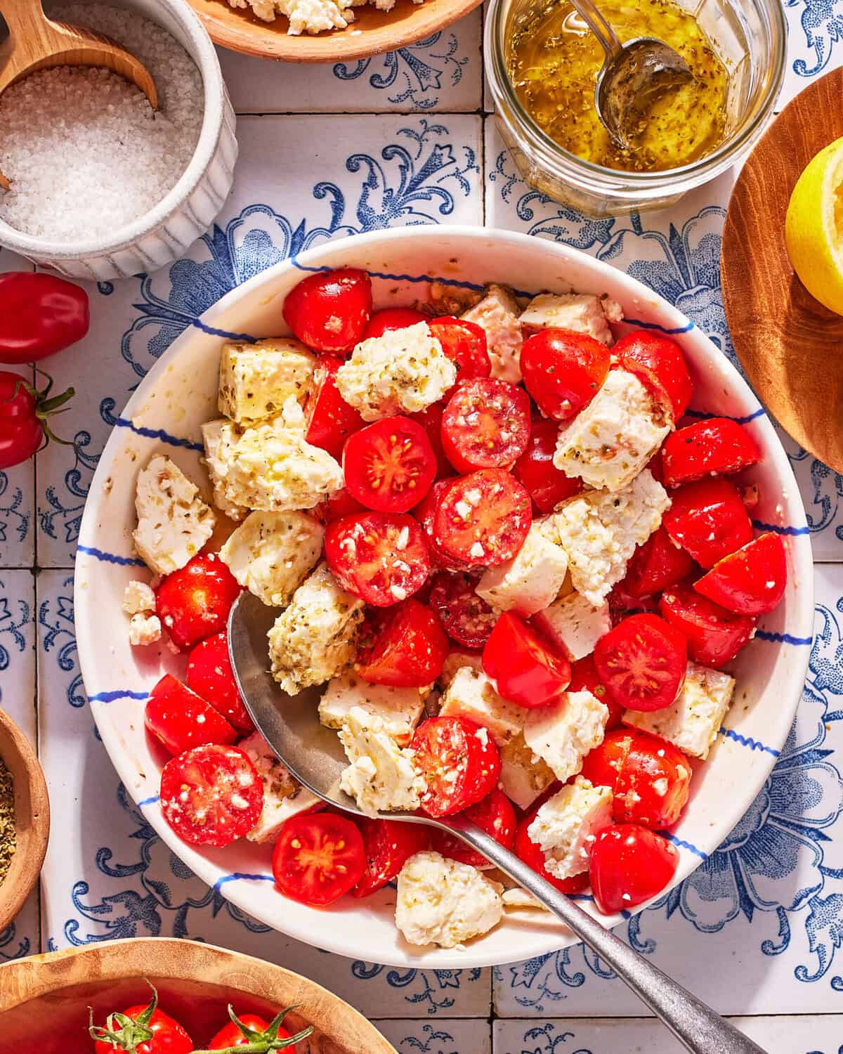 A bowl of cherry tomatoes and feta cheese chunks sits on a patterned tile surface, surrounded by small bowls of salt, herbs, dressing, and fresh tomatoes. A spoon rests in the tomato and feta salad.