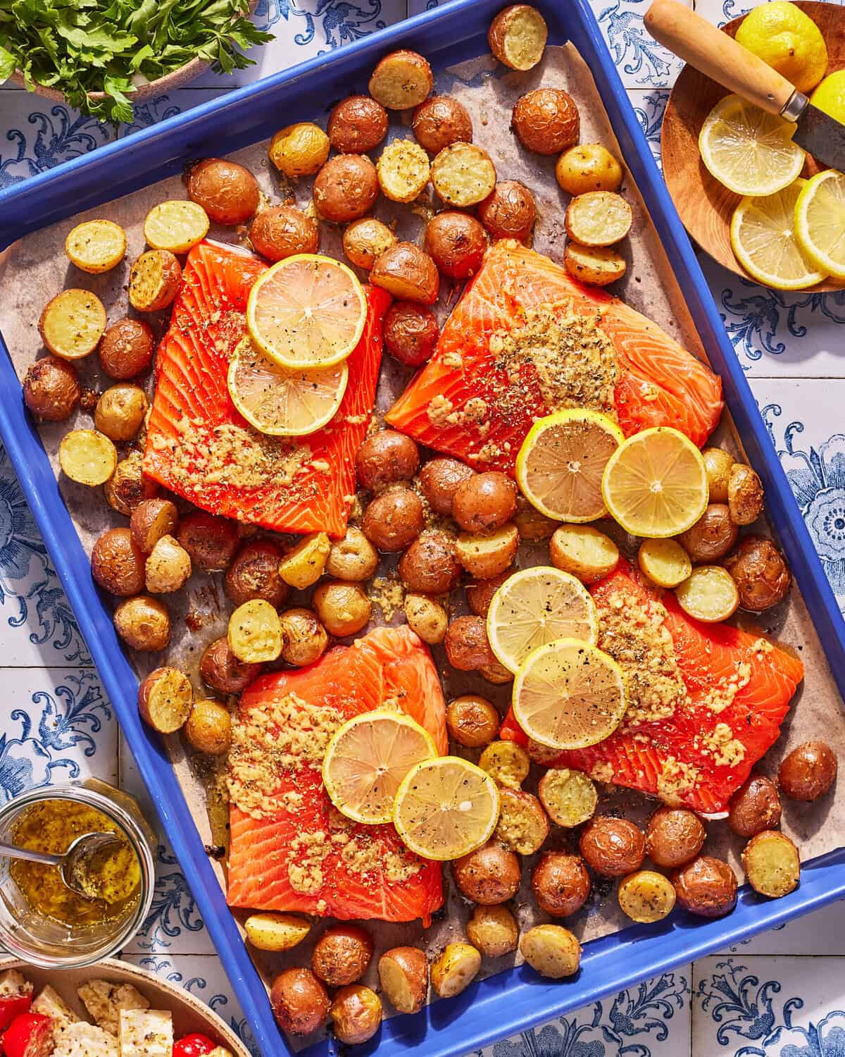 Baked salmon filets topped with lemon slices and seasoning, surrounded by roasted baby potatoes on a blue baking tray, with herbs, a bowl of feta, and a sauce jar nearby on a patterned table.
