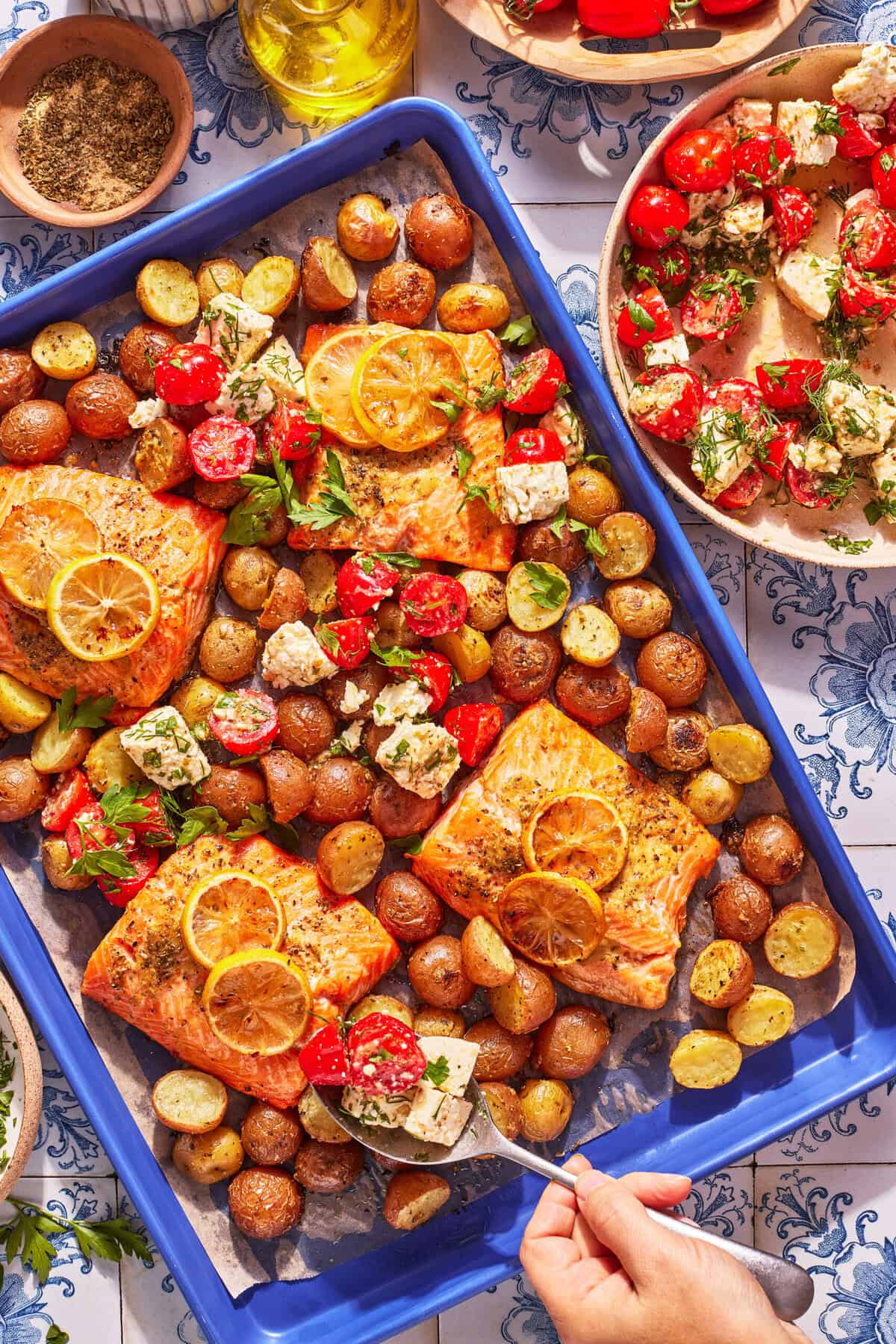 Baked salmon fillets with lemon slices, baby potatoes, cherry tomatoes, and cubes of feta cheese on a blue baking tray, with a hand serving some on a plate.