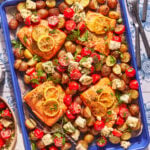 A blue baking tray with roasted salmon fillets topped with lemon slices, surrounded by roasted potatoes, cherry tomatoes, herbs, and cubes of feta cheese on a decorated table.