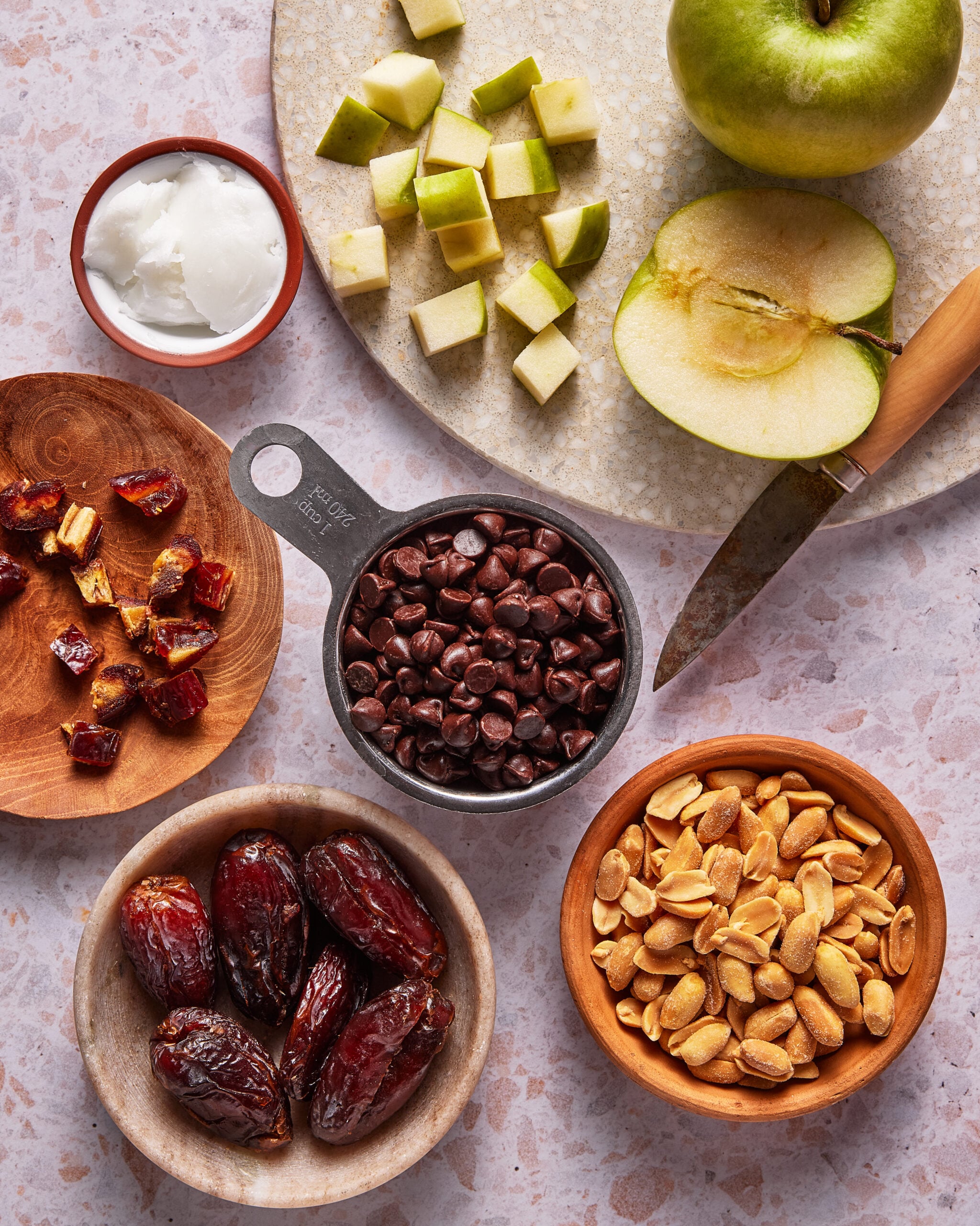 Bowls of dates, chopped dates, peanuts, chocolate chips, Greek yogurt, and diced green apple on a countertop, with a knife and halved green apple beside them.