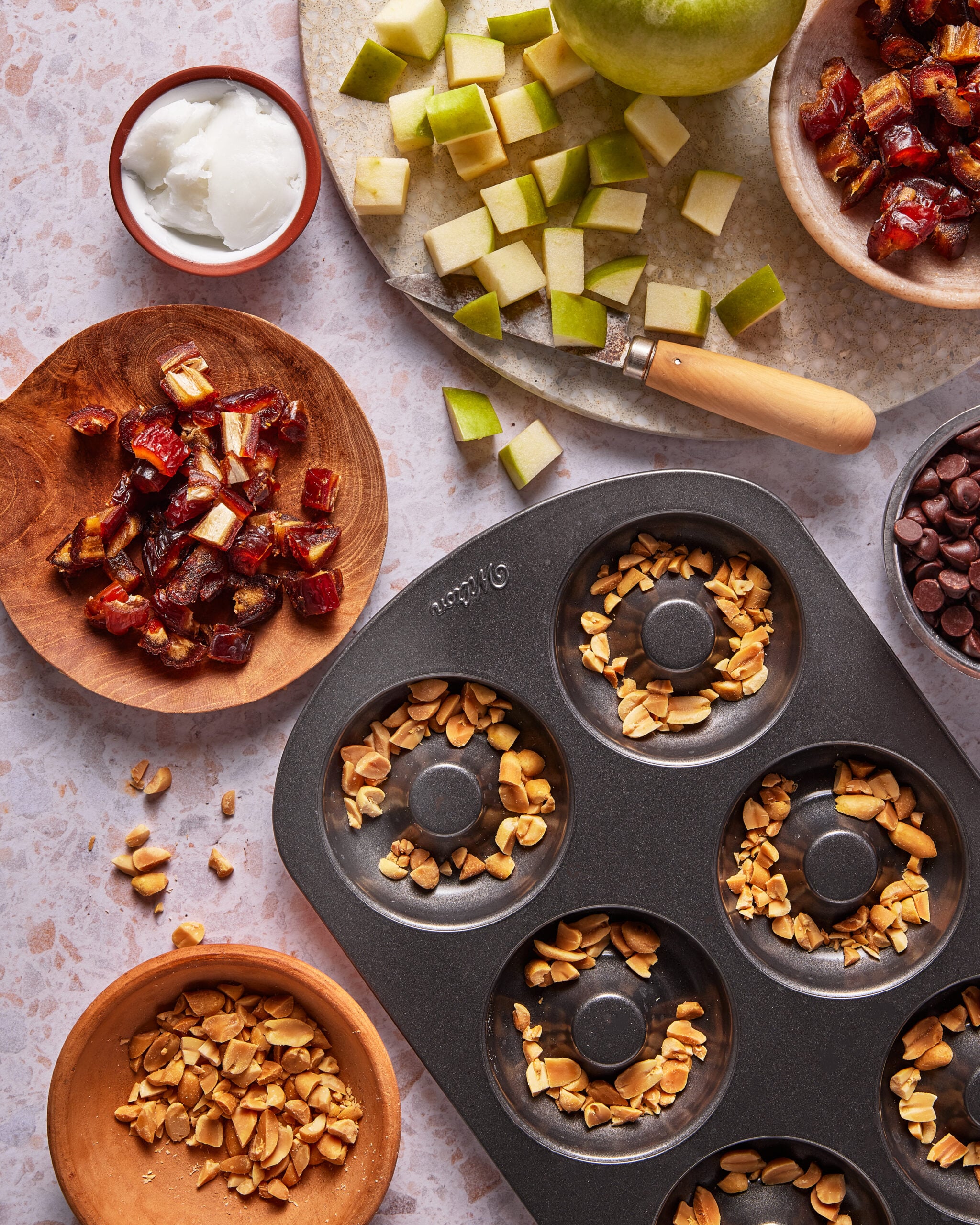 A donut pan with chopped peanuts, surrounded by bowls of chopped dates, apple pieces, chocolate chips, yogurt, and a cutting board with a knife. Ingredients are neatly arranged on a light countertop.
