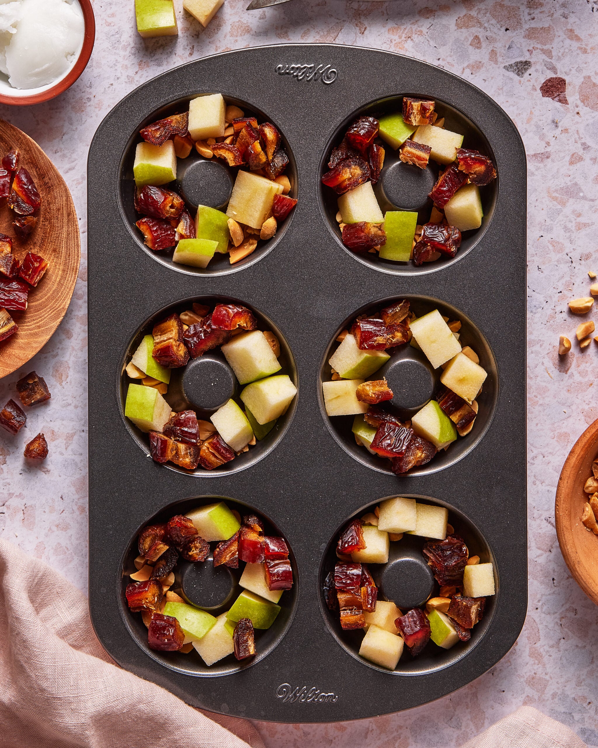 A muffin tin with six wells, each filled with chopped dates, green and red apple pieces, and nuts, sits on a speckled countertop surrounded by small bowls of ingredients.