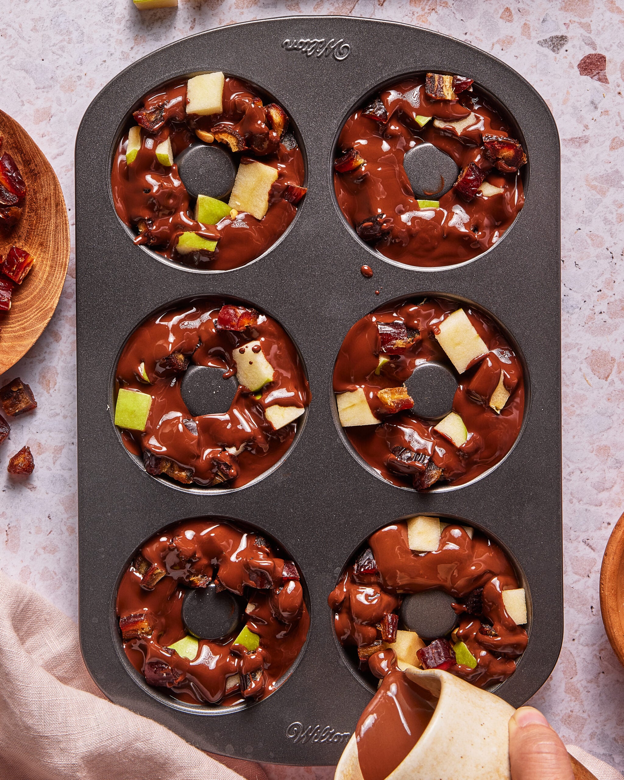 A donut pan with six cavities filled with melted chocolate, apple chunks, and dried fruit pieces. A hand pours more chocolate into the pan. The pan rests on a light, speckled surface.