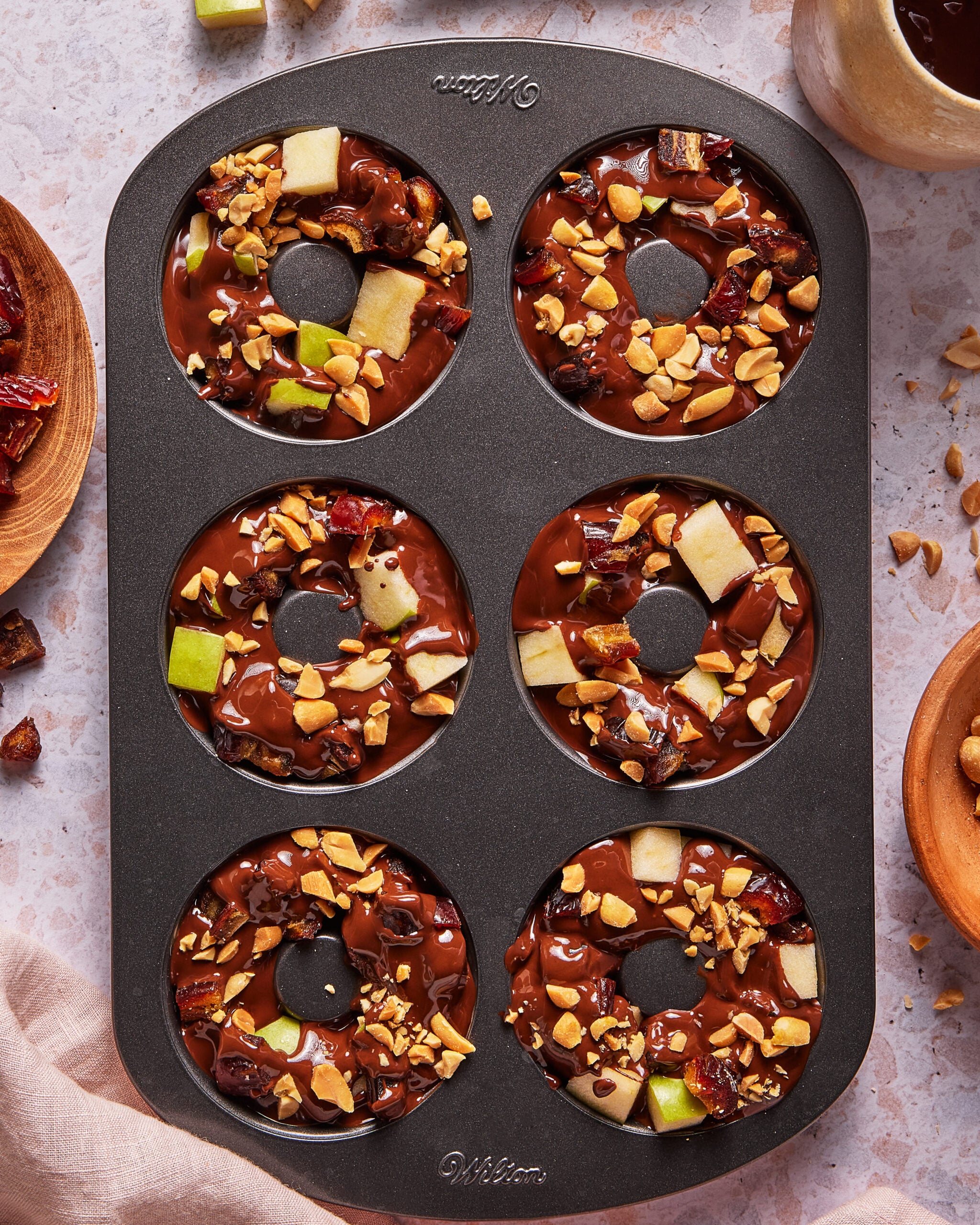 A nonstick donut pan holds six chocolate-coated donuts topped with chopped peanuts, green apple pieces, and dried fruit pieces. A cup of coffee and bowls of ingredients are nearby on a light countertop.