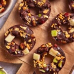 Chocolate-covered apple rings topped with chopped peanuts, dates, and a sprinkle of sea salt, arranged on a piece of parchment paper with a bowl of additional toppings nearby.