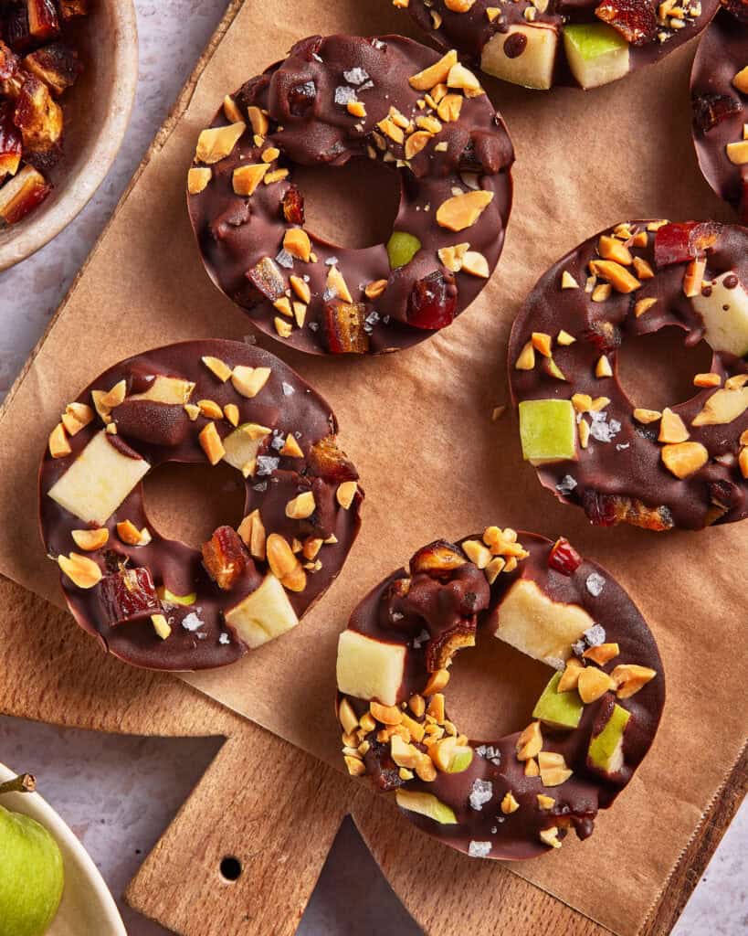 Chocolate-covered apple rings topped with chopped peanuts, dates, and a sprinkle of sea salt, arranged on a piece of parchment paper with a bowl of additional toppings nearby.