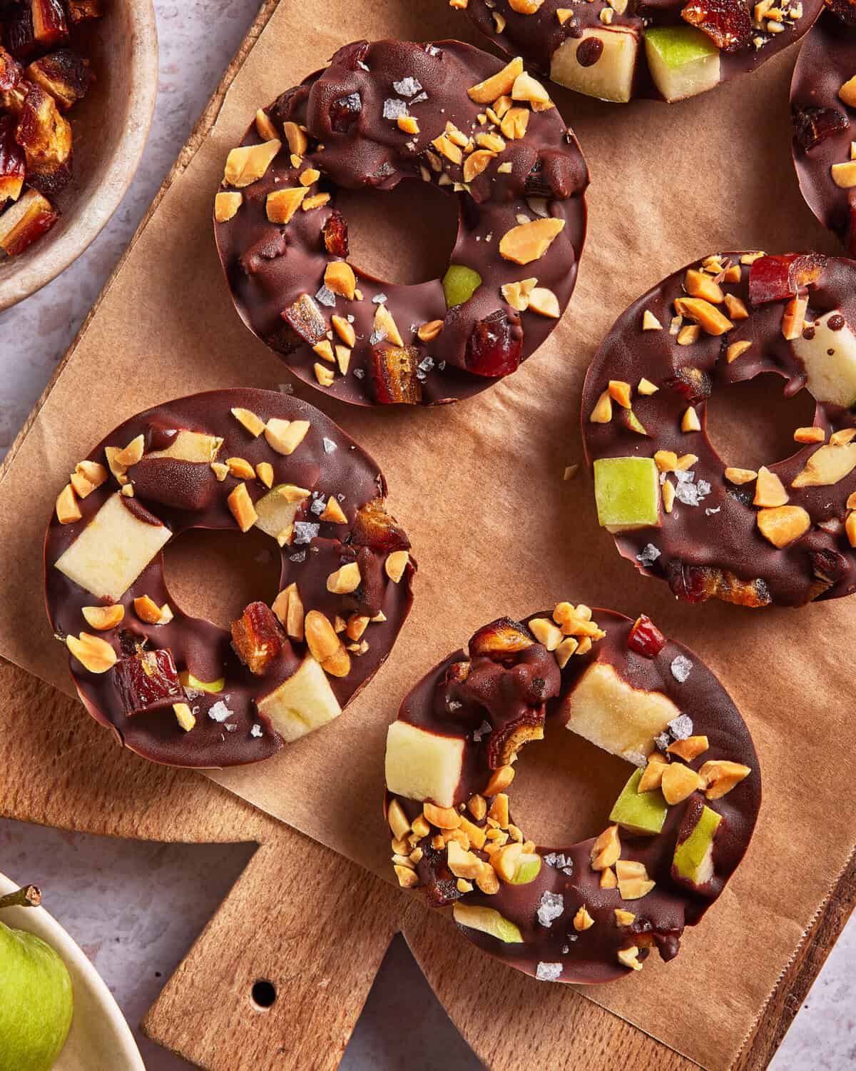 Chocolate-covered apple rings topped with chopped peanuts, dates, and a sprinkle of sea salt, arranged on a piece of parchment paper with a bowl of additional toppings nearby.