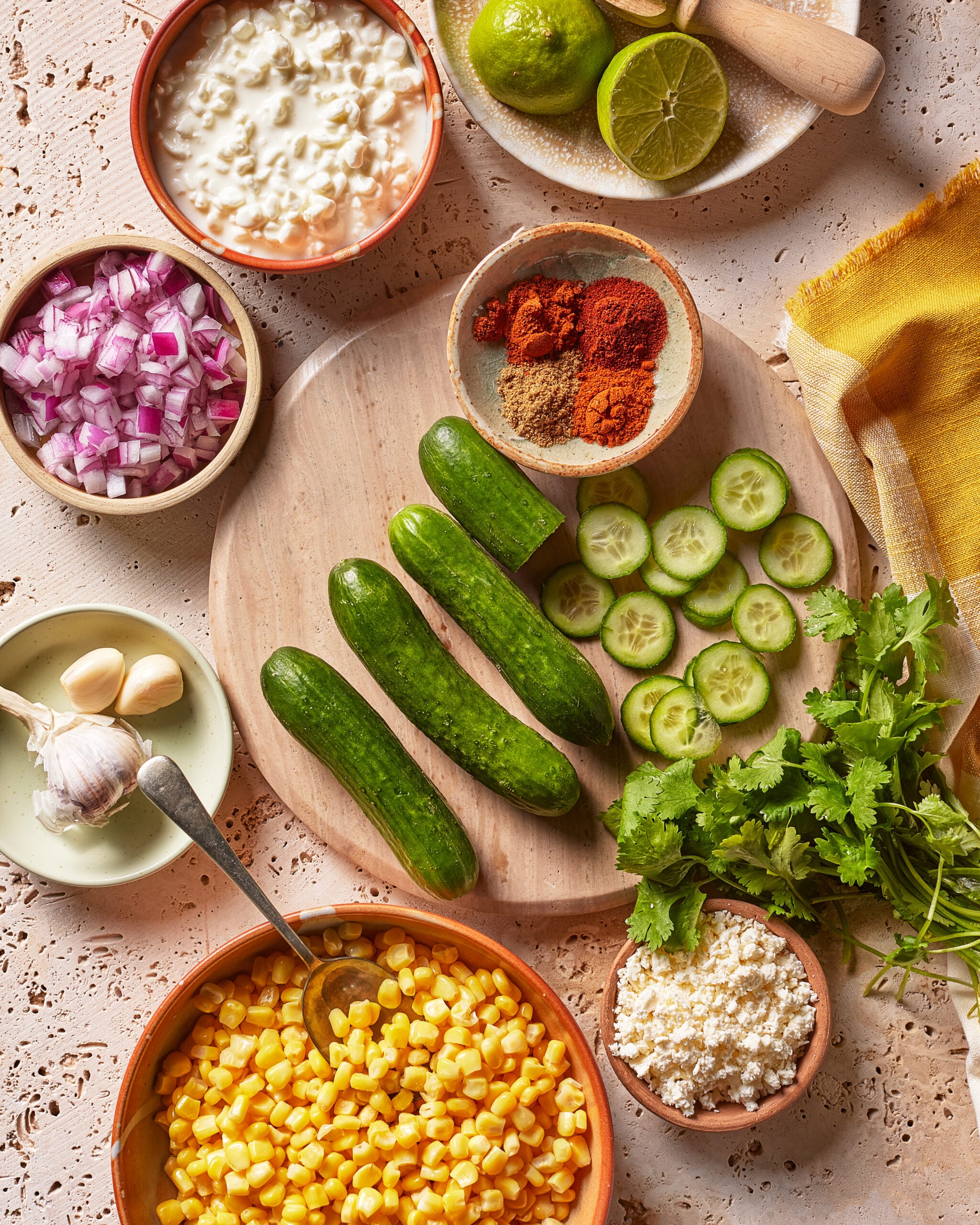 Fresh ingredients on a table: whole and sliced cucumbers, corn, red onion, limes, garlic, cilantro, cottage cheese, crumbled cheese, and small bowls of spices on a light textured surface.