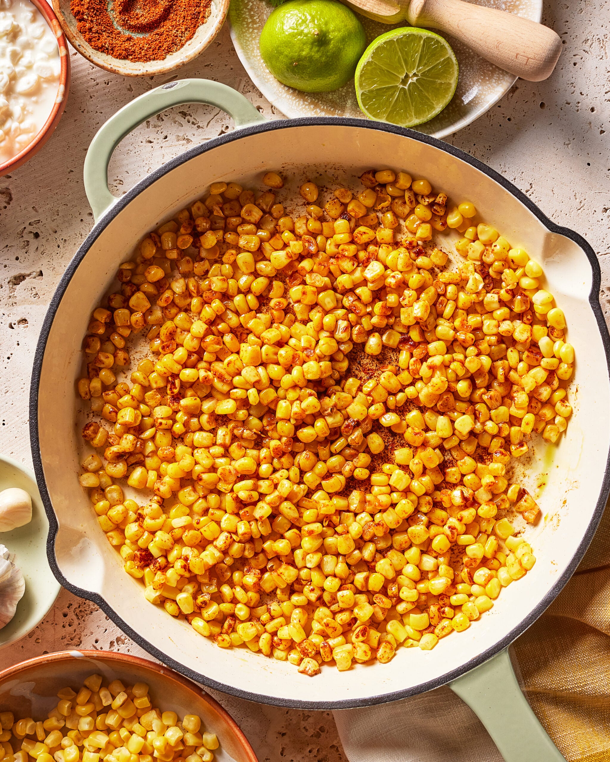 Cooked corn kernels sautéed with spices in a large white skillet, with lime halves, seasoning, and other ingredients visible around the pan on a textured countertop.