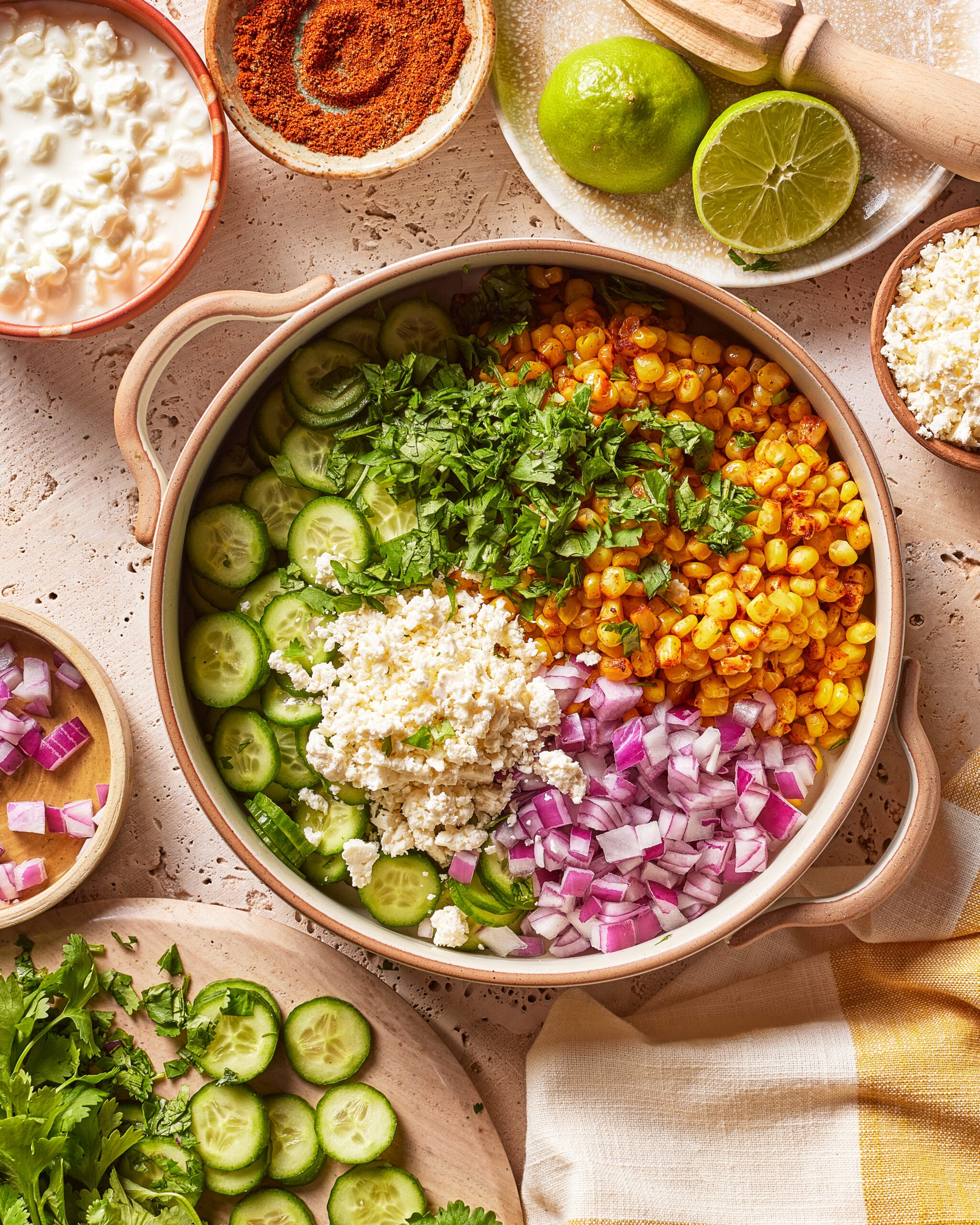 A bowl with sliced cucumbers, roasted corn, chopped red onion, cheese, and cilantro, surrounded by lime, spices, more cheese, cottage cheese, and fresh vegetables on a textured surface.