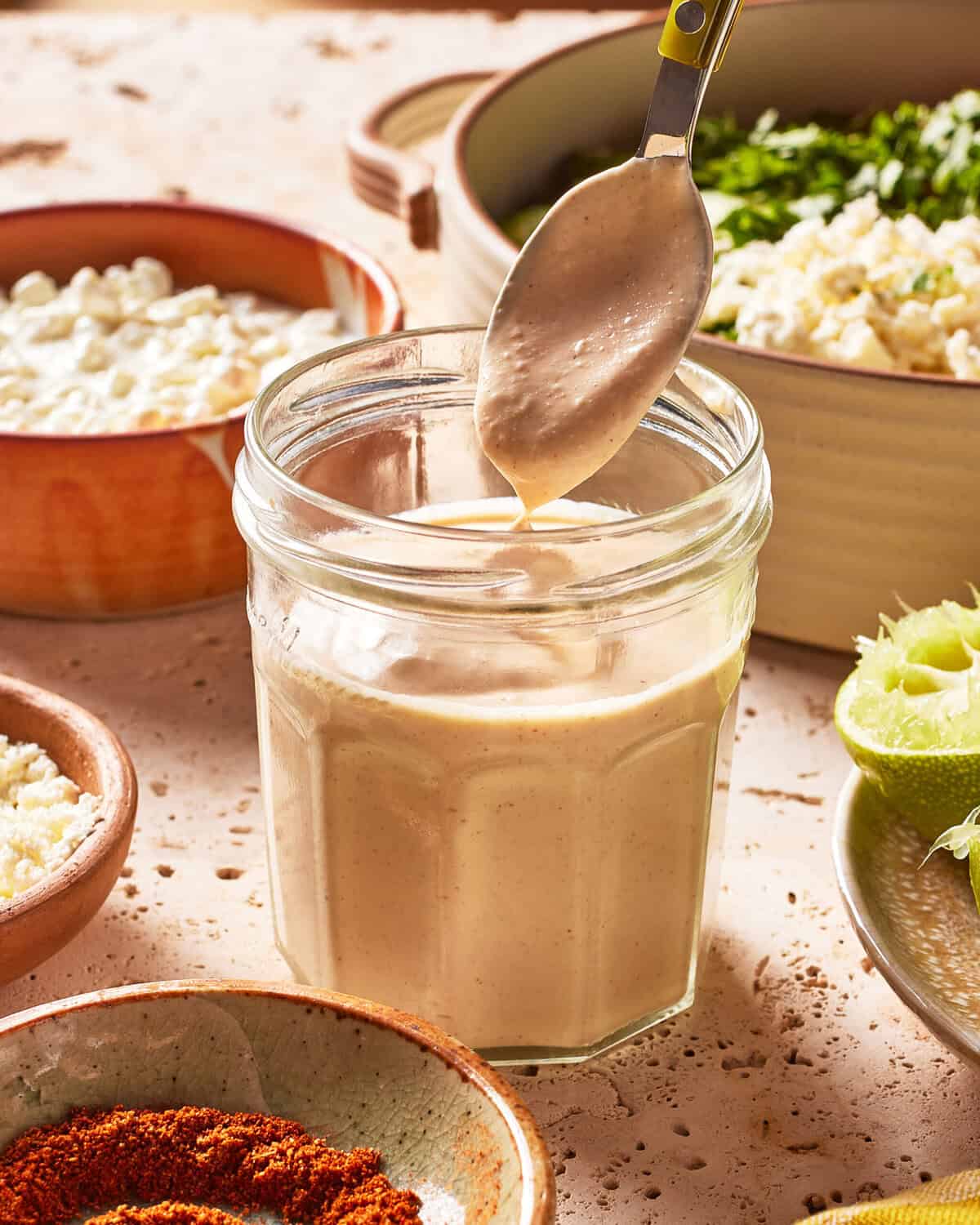 A spoon scoops creamy sauce from a glass jar, surrounded by bowls of spices, cheese, herbs, and a halved lime on a sunlit countertop.