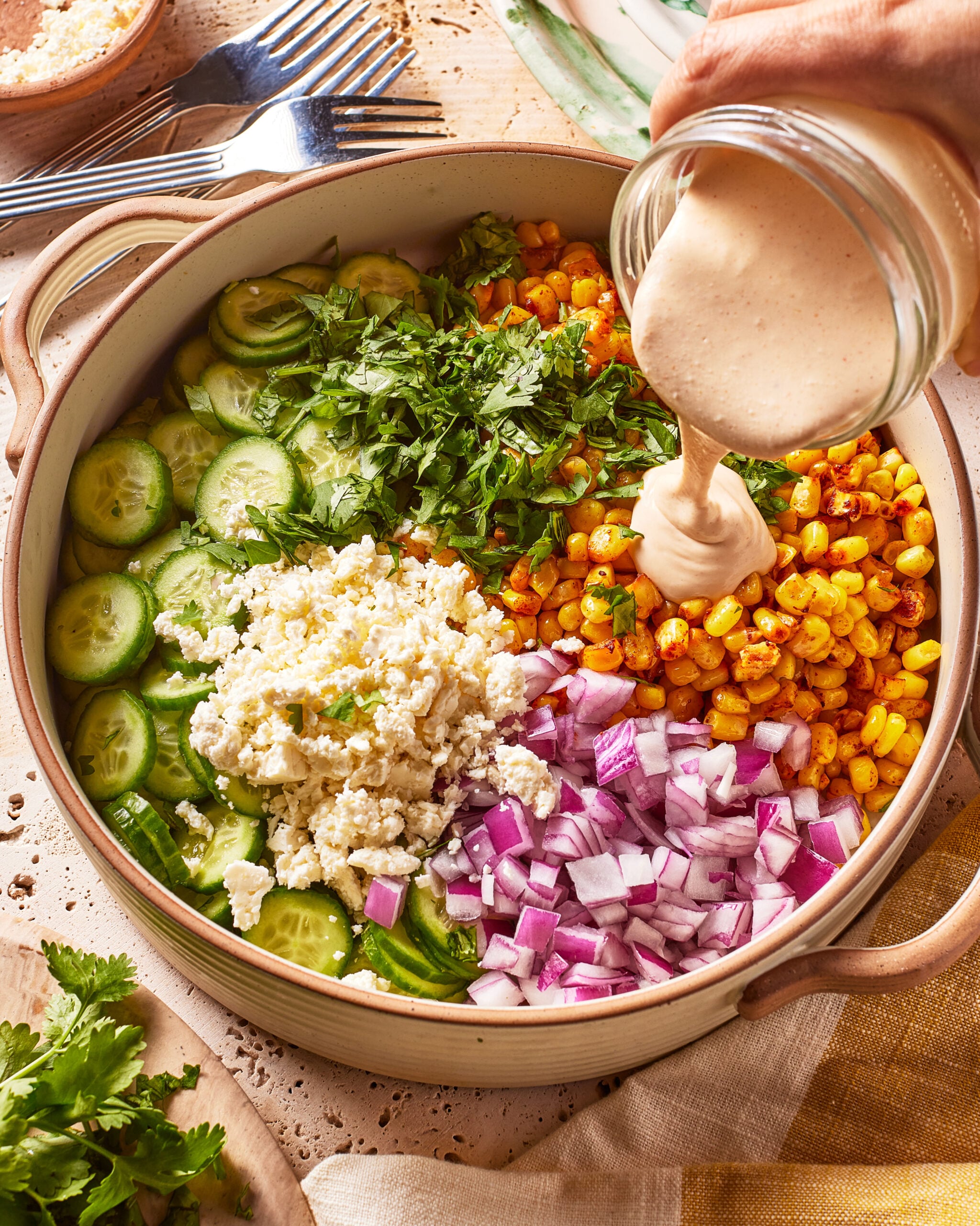 A bowl with sliced cucumbers, corn, red onion, cilantro, and cheese, while a hand pours creamy dressing over the salad. Forks and ingredients are visible in the background.