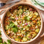 A bowl of salad with sliced cucumbers, corn, crumbled cheese, chopped red onion, and cilantro, with forks and a plate nearby on a rustic surface.