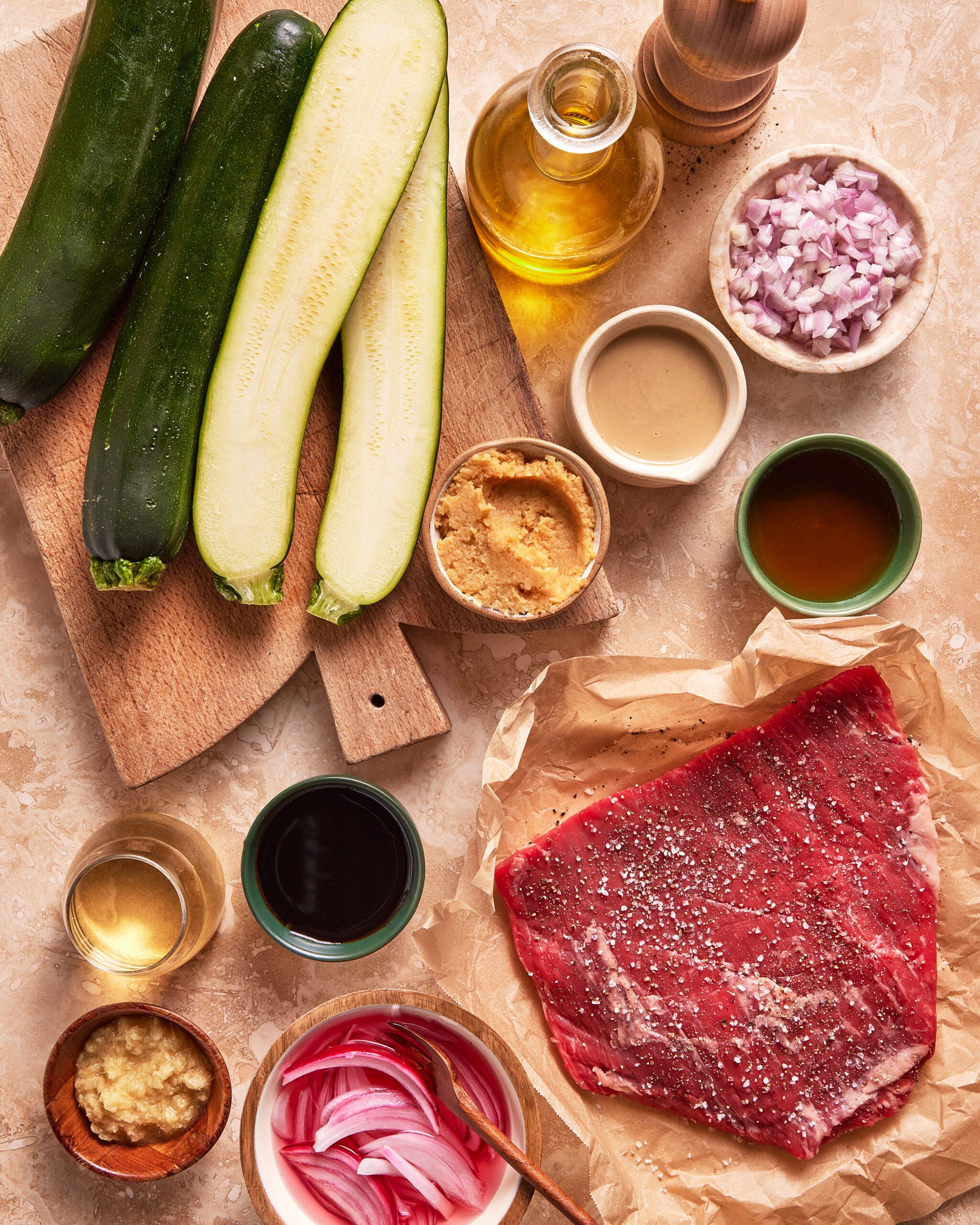 Raw steak with pepper and salt, whole and halved zucchinis, chopped onions, olive oil, vinegar, sauces, and seasonings arranged on a beige surface with a wooden cutting board and a pepper grinder.