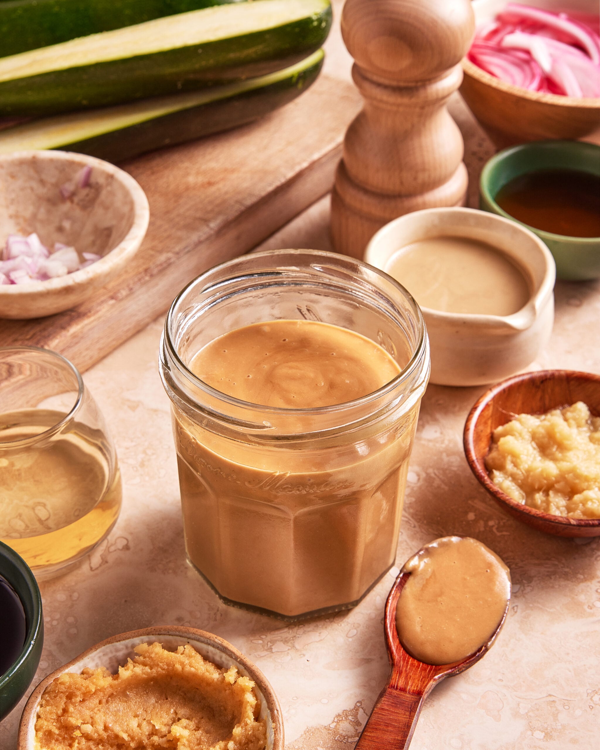 A jar of creamy dressing sits on a counter surrounded by bowls of ingredients like garlic, miso, vinegar, and tahini, with a wooden spoon holding some dressing and fresh vegetables in the background.