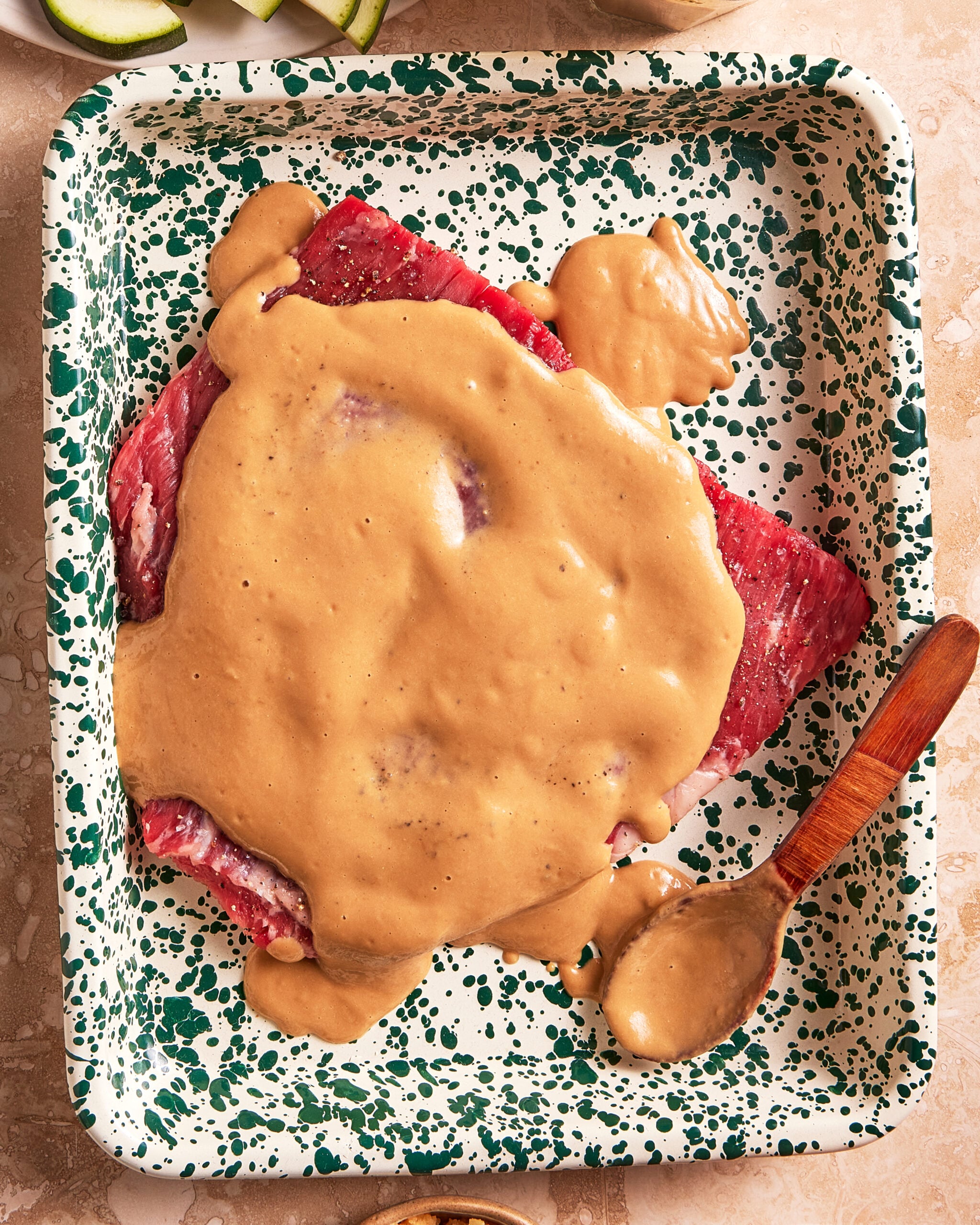 Raw steak in a green-speckled baking dish, covered with a thick, creamy brown sauce. A wooden spoon with some sauce rests on the edge of the dish.