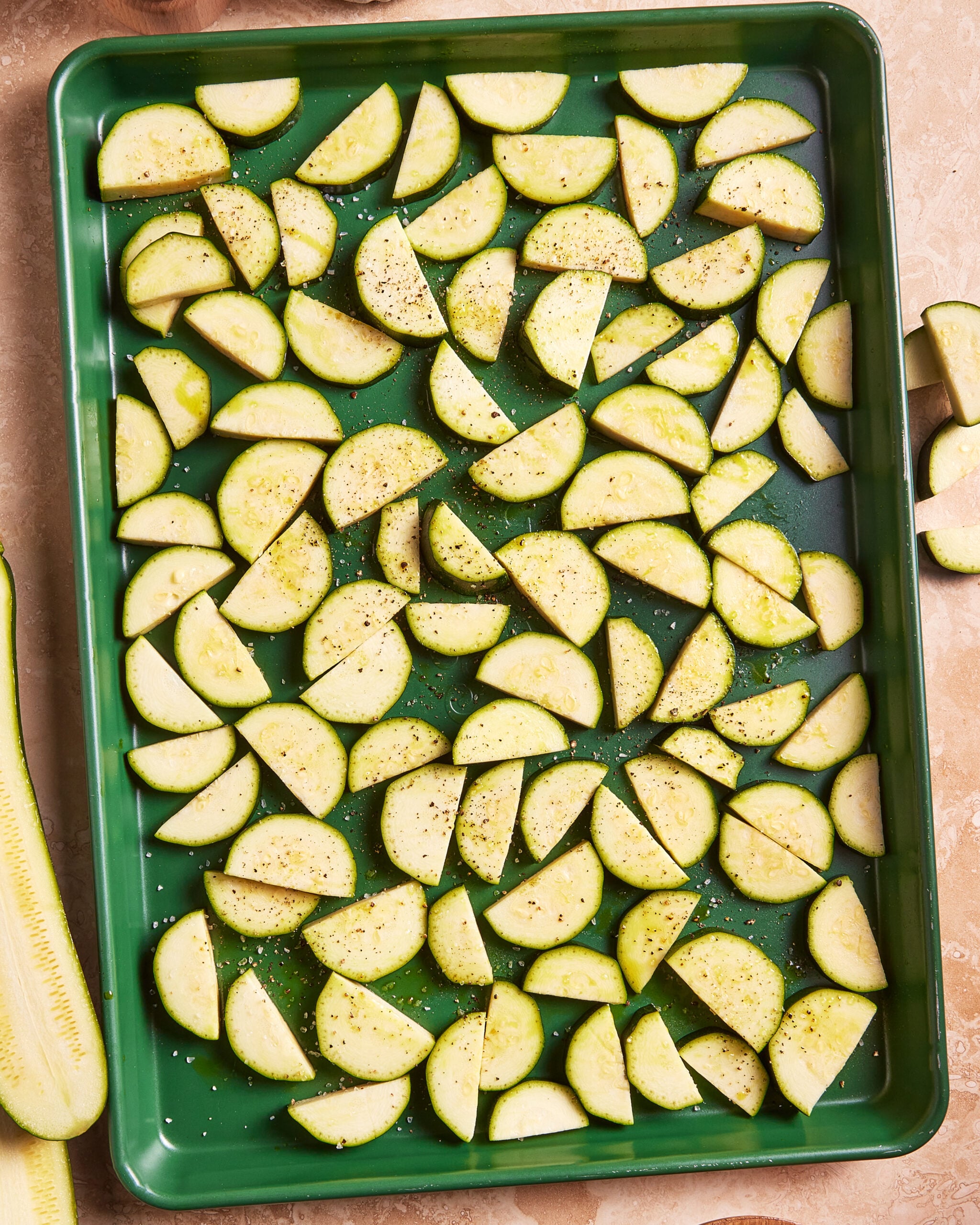 Sliced zucchini arranged on a green baking sheet, sprinkled with salt and pepper, ready to be roasted. The zucchini pieces are evenly spread out on the tray.