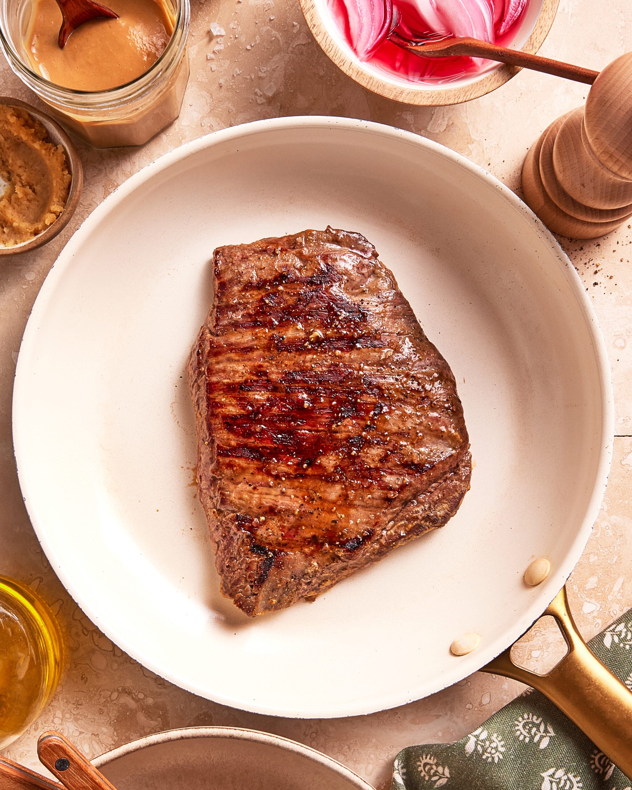 A grilled steak sits in a white skillet surrounded by condiments, a pepper grinder, and a green patterned napkin on a beige surface.