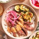 A bowl of white rice topped with sliced steak, grilled zucchini, pickled red onions, sesame seeds, and a creamy sauce. Bowls of rice, pickled onions, sesame seeds, and zucchini are nearby. Fork and napkin on the side.