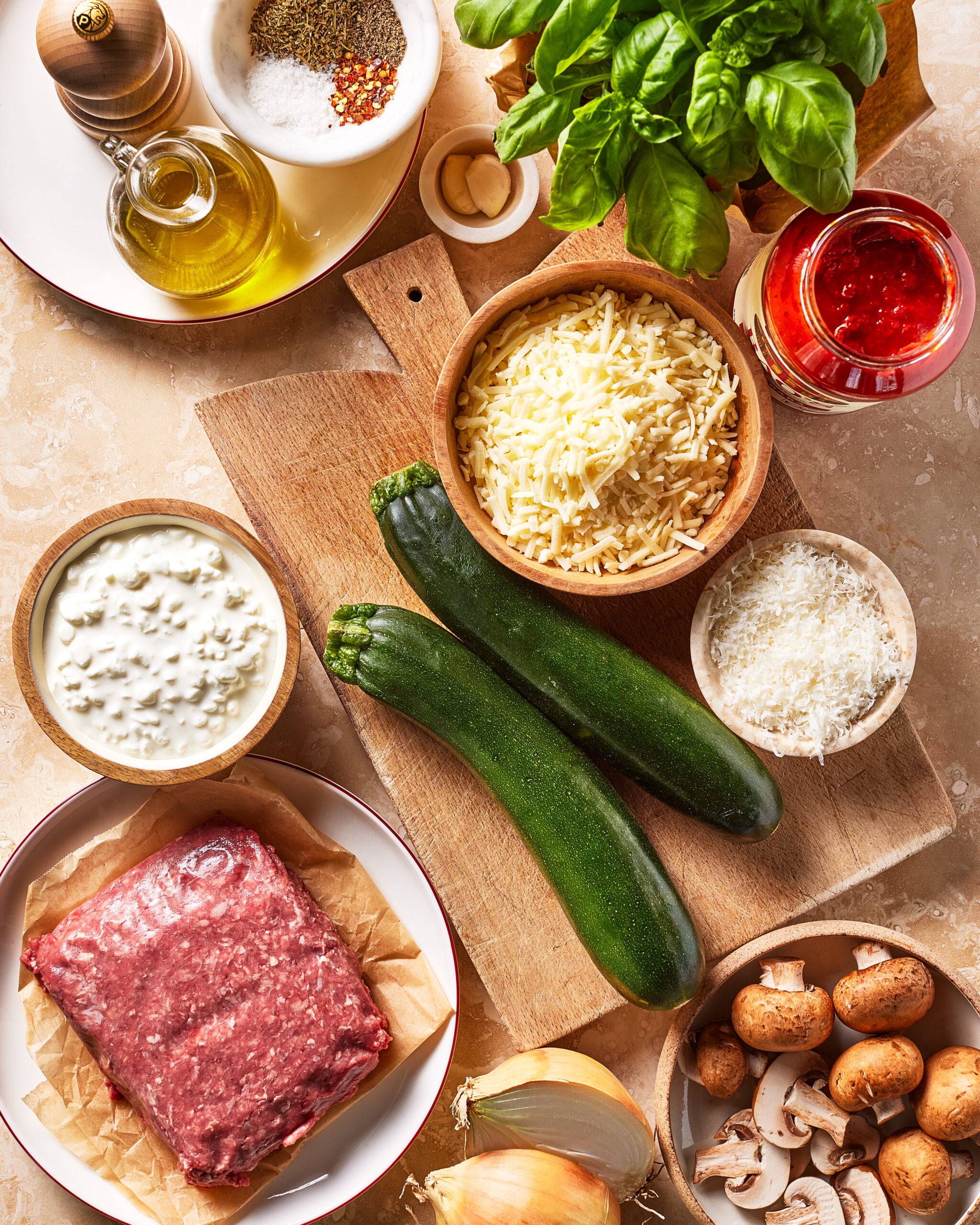 Ingredients for a recipe are laid out, including ground beef, zucchini, cheese, mushrooms, an onion, basil, olive oil, garlic, tomato sauce, and seasonings on a wooden board and countertop.