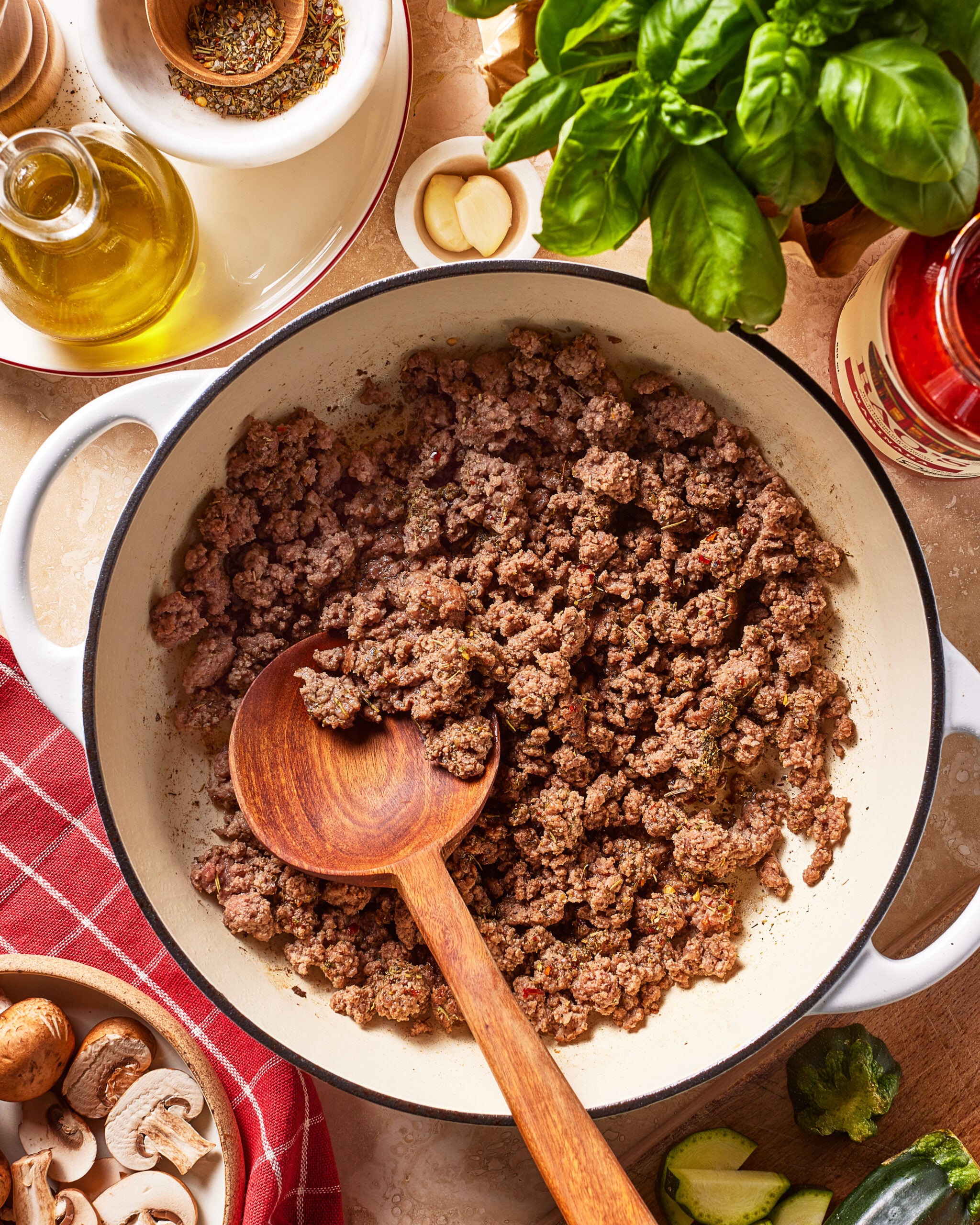 A wooden spoon stirs browned ground beef in a white pot, surrounded by fresh basil, olive oil, garlic, a red towel, mushrooms, zucchini, and seasonings on a kitchen counter.