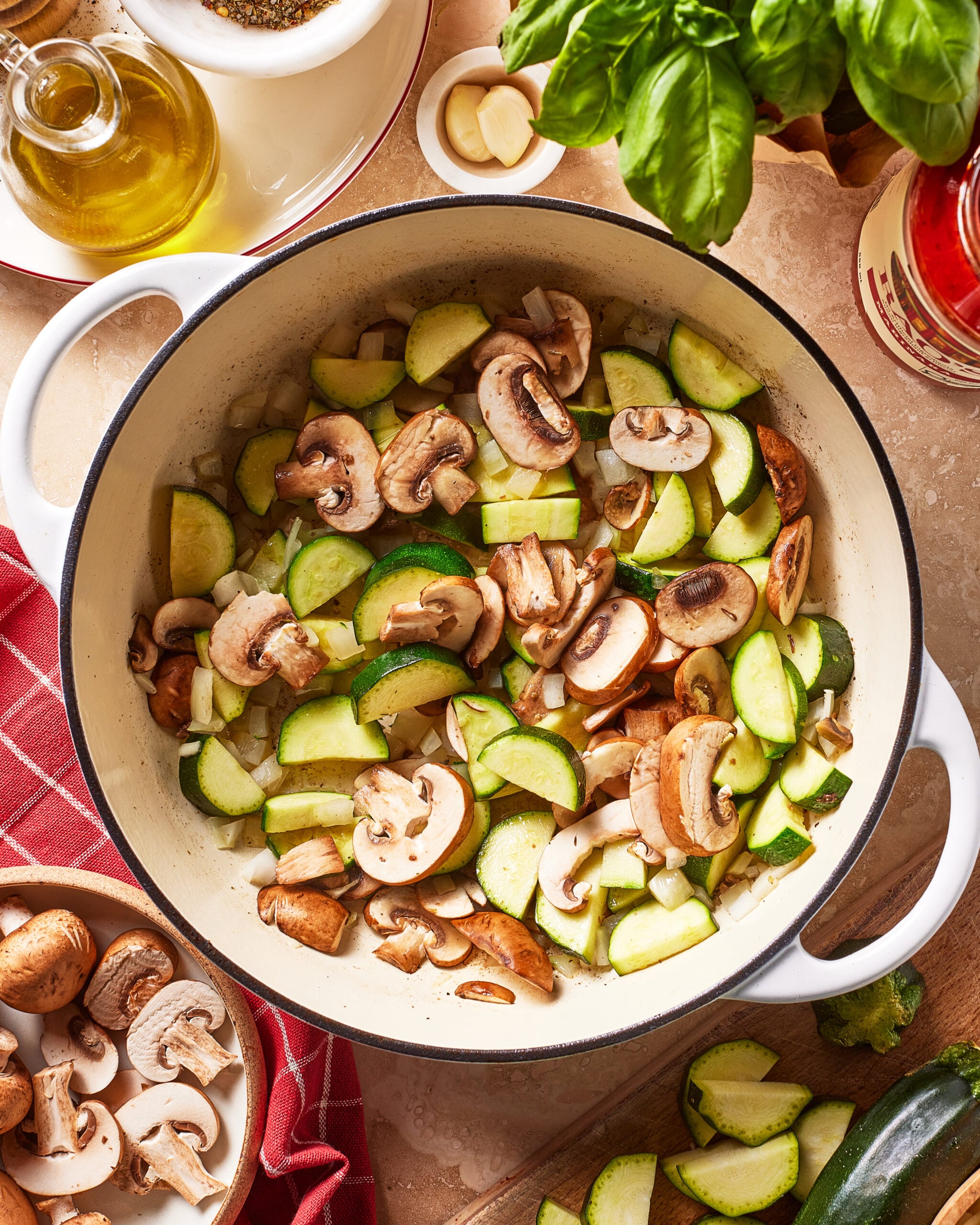 Sliced mushrooms and zucchini cooking in a white pot, surrounded by fresh basil, olive oil, a red towel, garlic, and a small bowl of mushrooms on a kitchen counter.