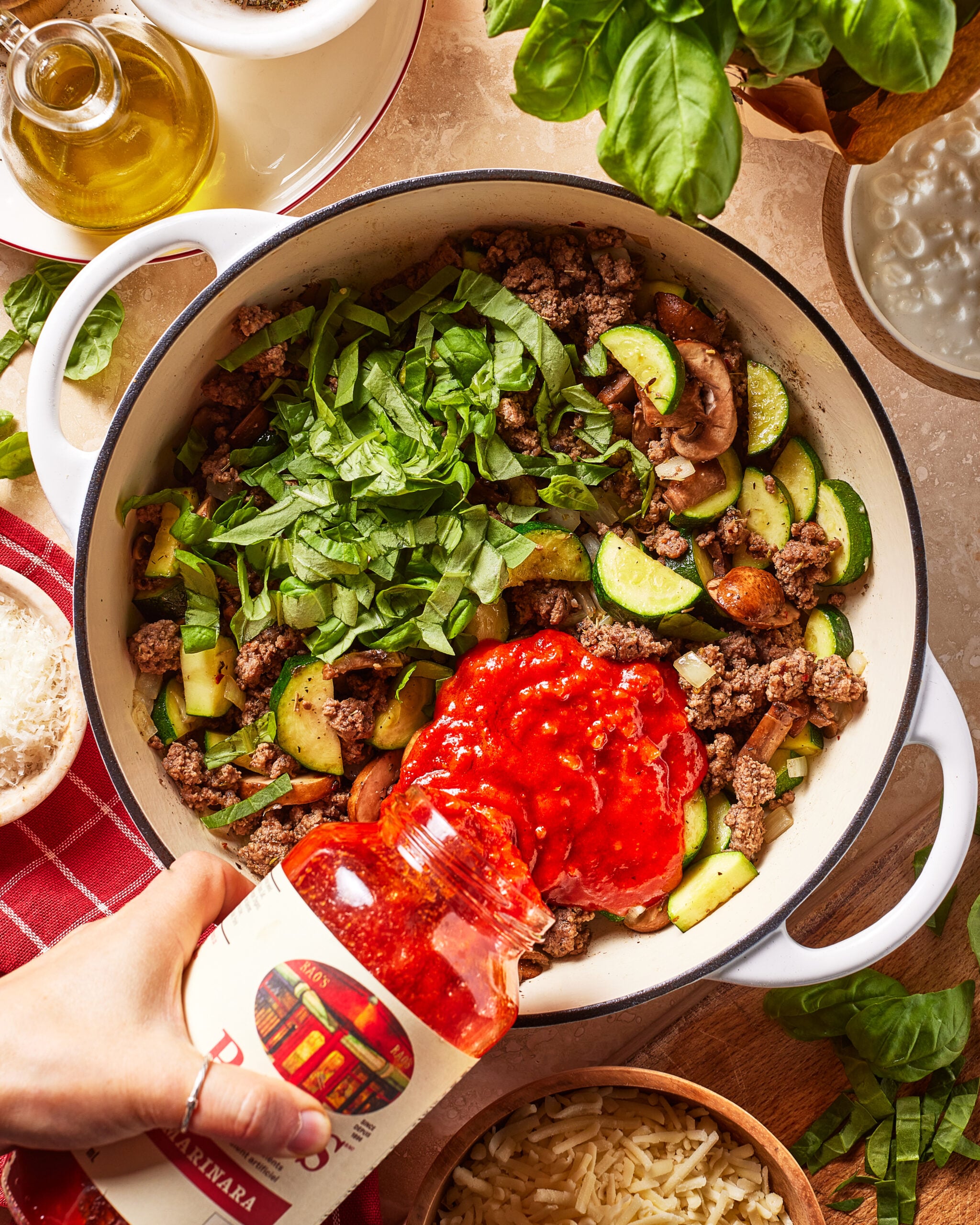 A hand pours jarred marinara sauce into a pot with ground beef, chopped spinach, zucchini, and mushrooms. Fresh basil, olive oil, a red towel, and a bowl of shredded cheese are nearby.