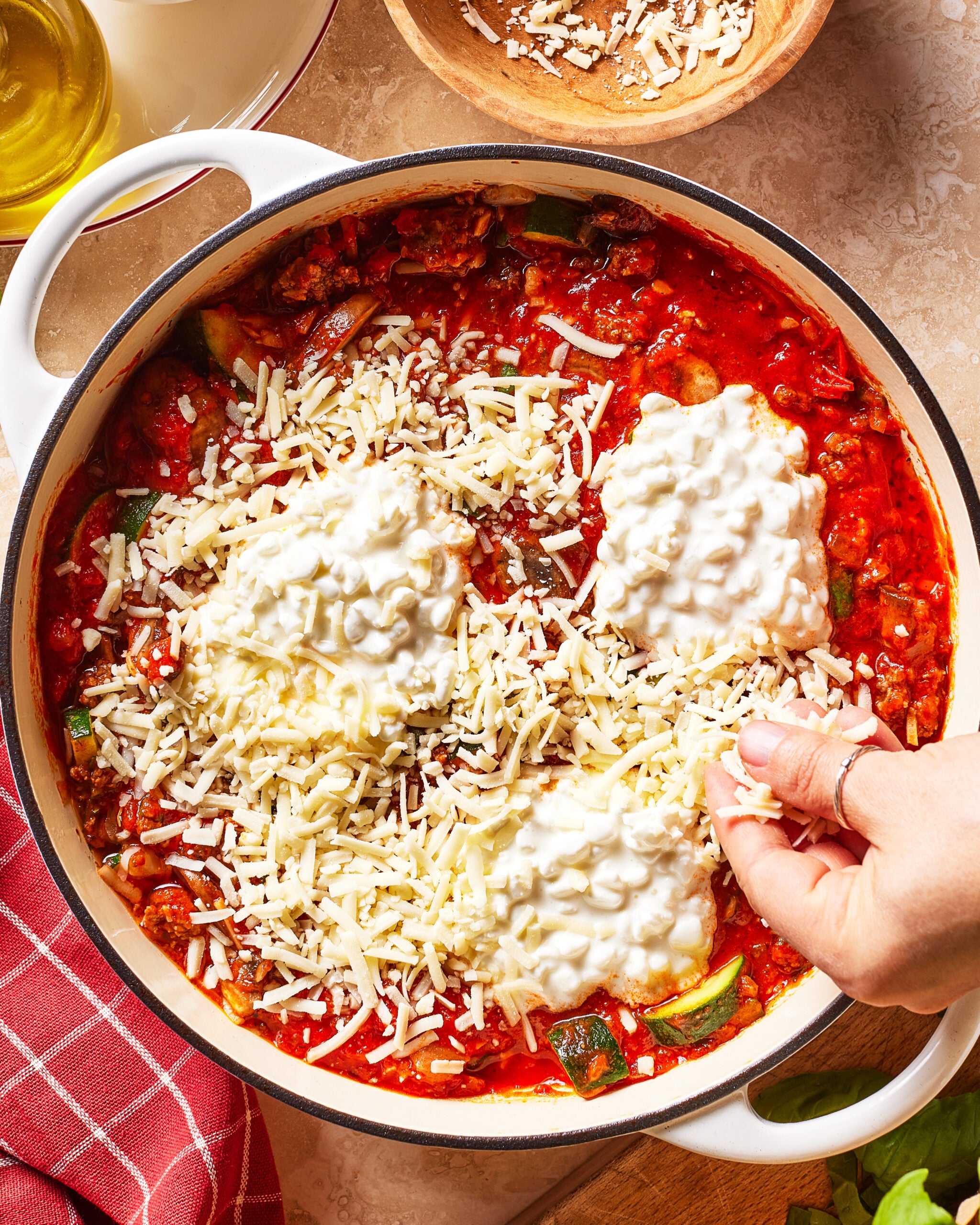 A hand sprinkles shredded cheese over a pot of red pasta sauce topped with cottage cheese. A bowl of grated cheese and a towel are nearby on the countertop.