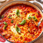 A baked skillet of cheesy lasagna with tomato sauce, garnished with fresh basil leaves, sits on a table next to a red napkin and serving plates.