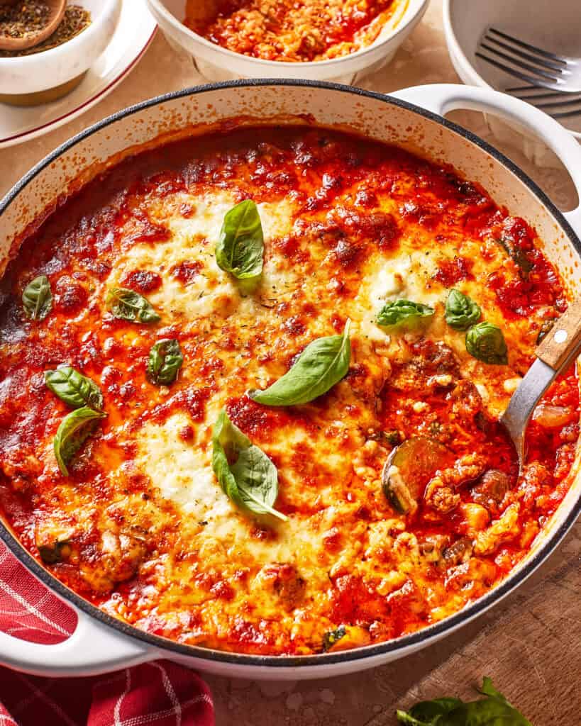 A baked skillet of cheesy lasagna with tomato sauce, garnished with fresh basil leaves, sits on a table next to a red napkin and serving plates.