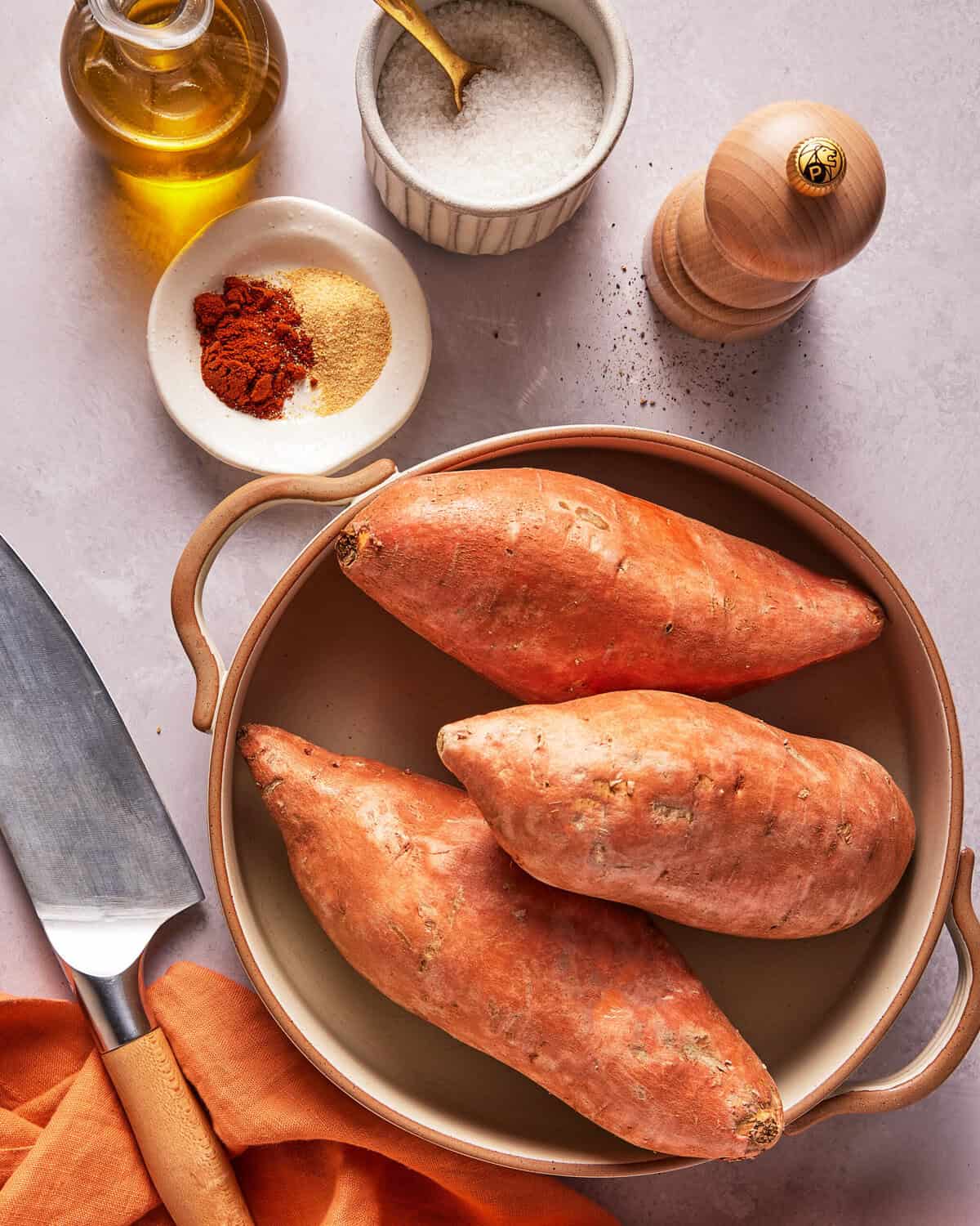 Three whole sweet potatoes in a round baking dish, surrounded by a knife, olive oil, dish of salt, pepper grinder, and a small bowl of paprika and garlic powder on a light surface.