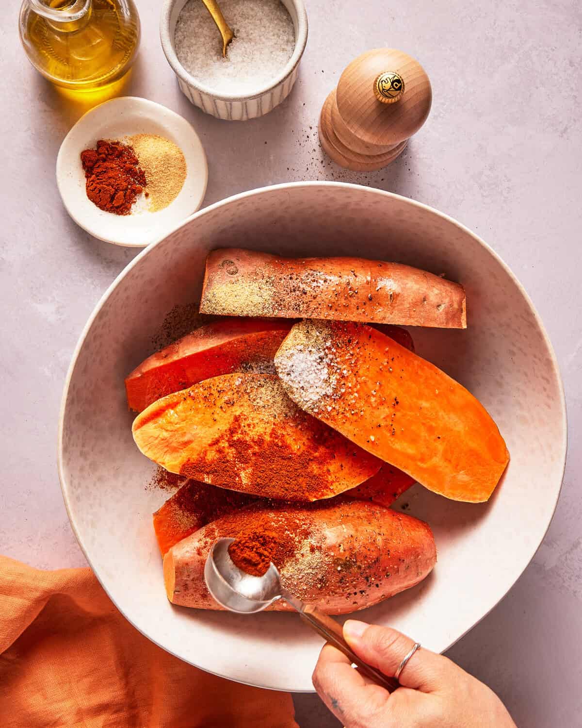 A hand sprinkles spices over sweet potato wedges in a bowl. Nearby are a small bowl of spices, salt, a pepper grinder, and a bottle of oil on a light countertop.