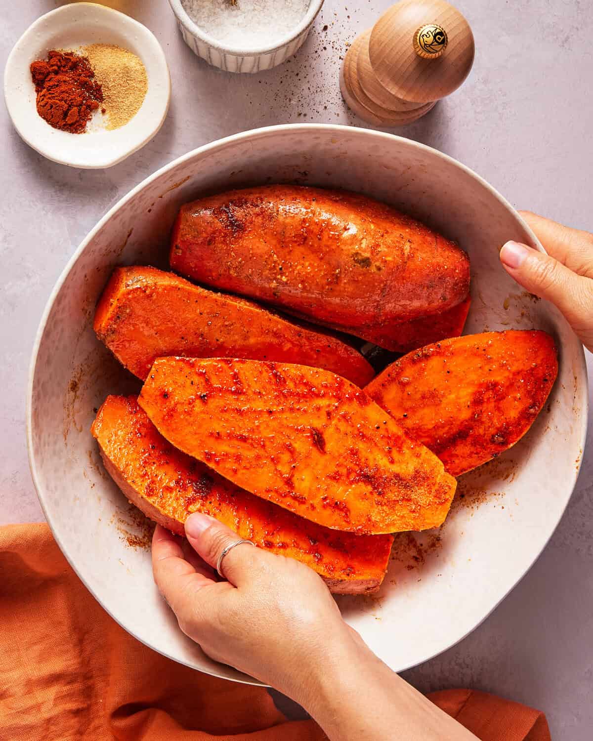 Hands holding a bowl of sliced sweet potatoes seasoned with spices. A small plate of spices, a pepper grinder, and a bowl of salt are nearby on a light surface. An orange cloth rests beside the bowl.