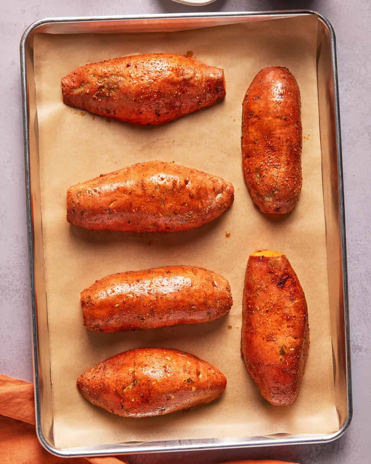 Six seasoned sweet potatoes on a parchment-lined baking sheet, ready for roasting.