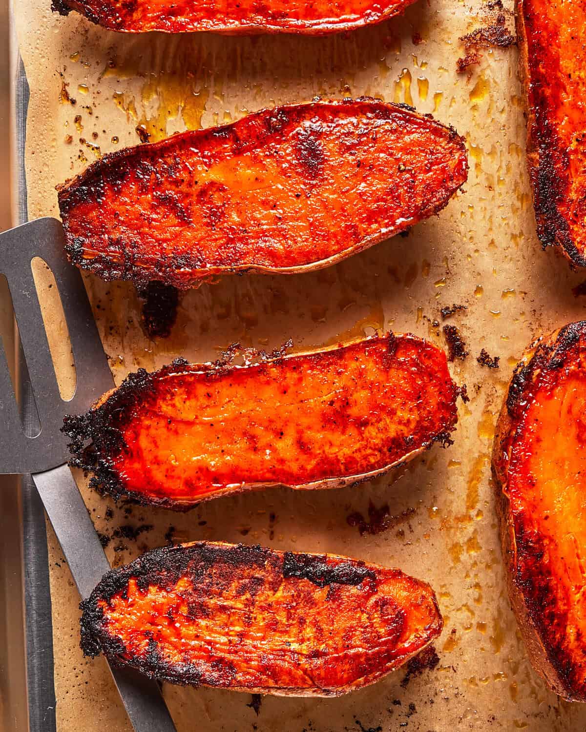 Roasted sweet potato halves with crispy, caramelized edges on a parchment-lined baking sheet, next to a metal spatula.