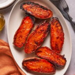 Six roasted sweet potato halves with charred edges, sprinkled with coarse salt, are arranged on an oval white plate. A fork, knife, cloth napkin, and condiment bowl are nearby.
