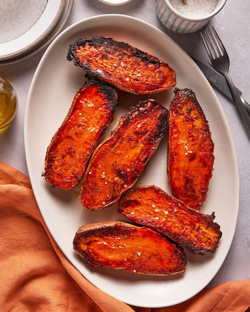Six roasted sweet potato halves with charred edges, sprinkled with coarse salt, are arranged on an oval white plate. A fork, knife, cloth napkin, and condiment bowl are nearby.
