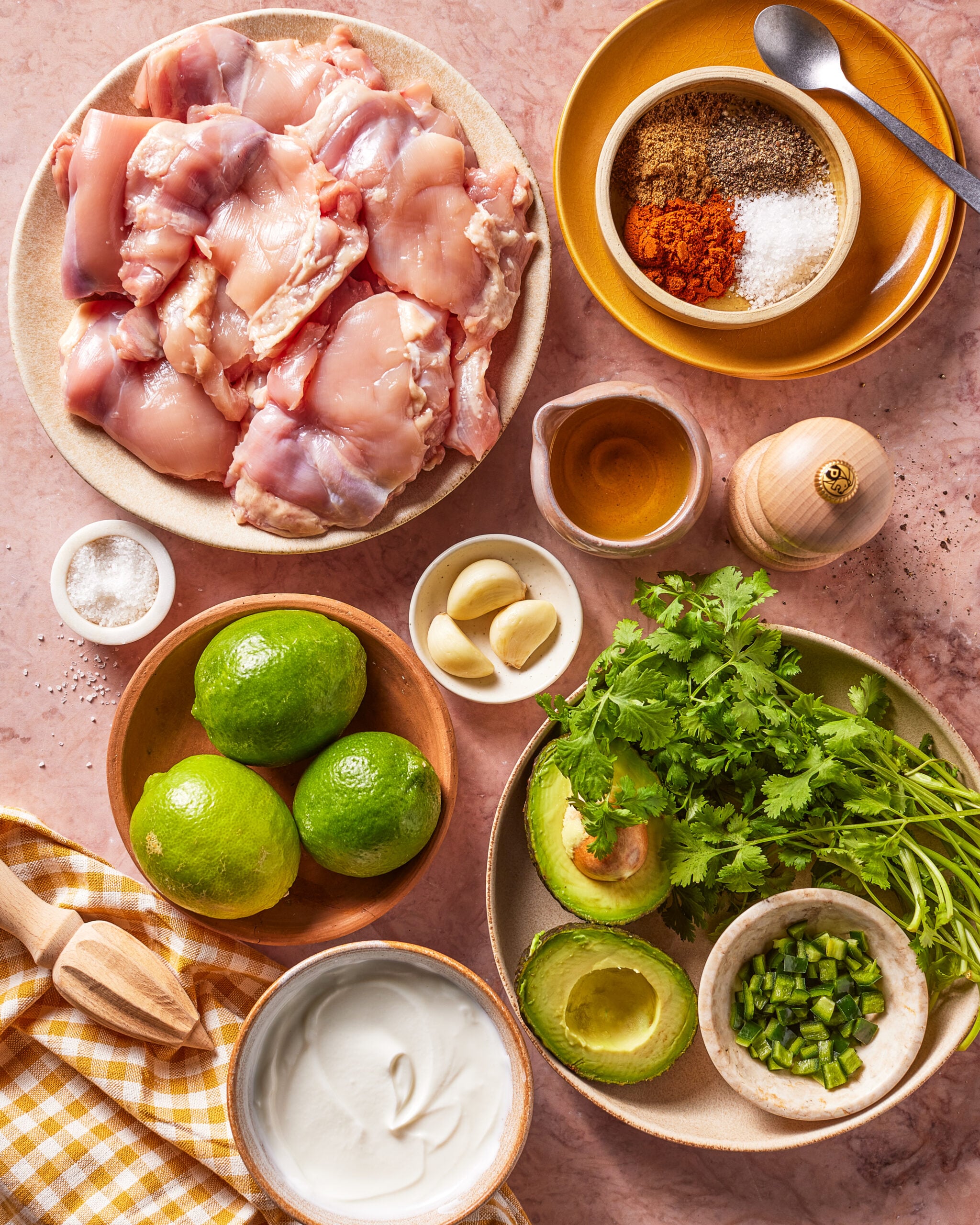 Raw chicken, spices, garlic, olive oil, limes, cilantro, avocado, jalapeño, salt, pepper, and a bowl of yogurt are arranged on a pink countertop with a yellow checkered napkin.
