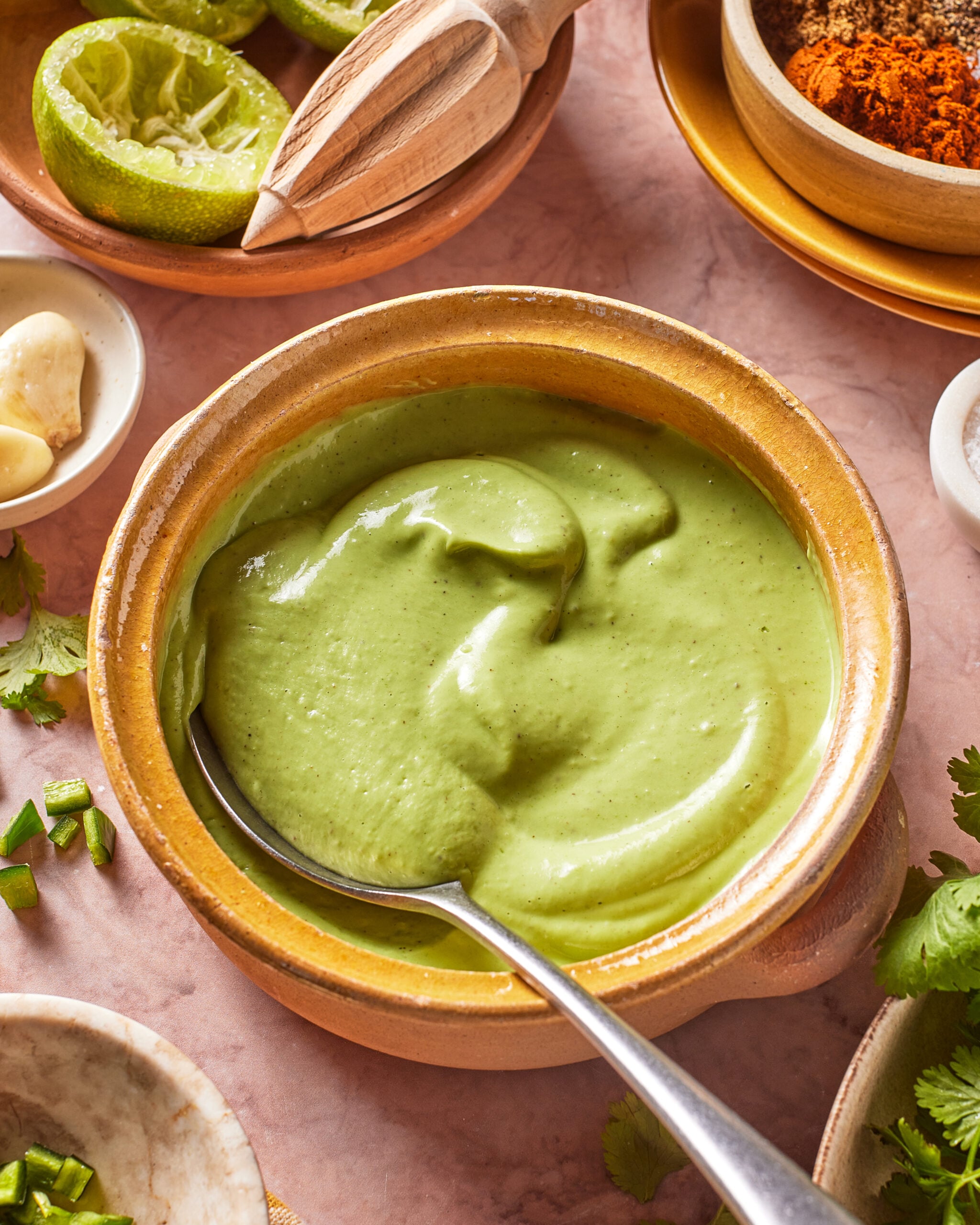 A bowl of creamy green sauce with a spoon, surrounded by lime halves, garlic, cilantro, spices, and jalapeno on a pink surface.