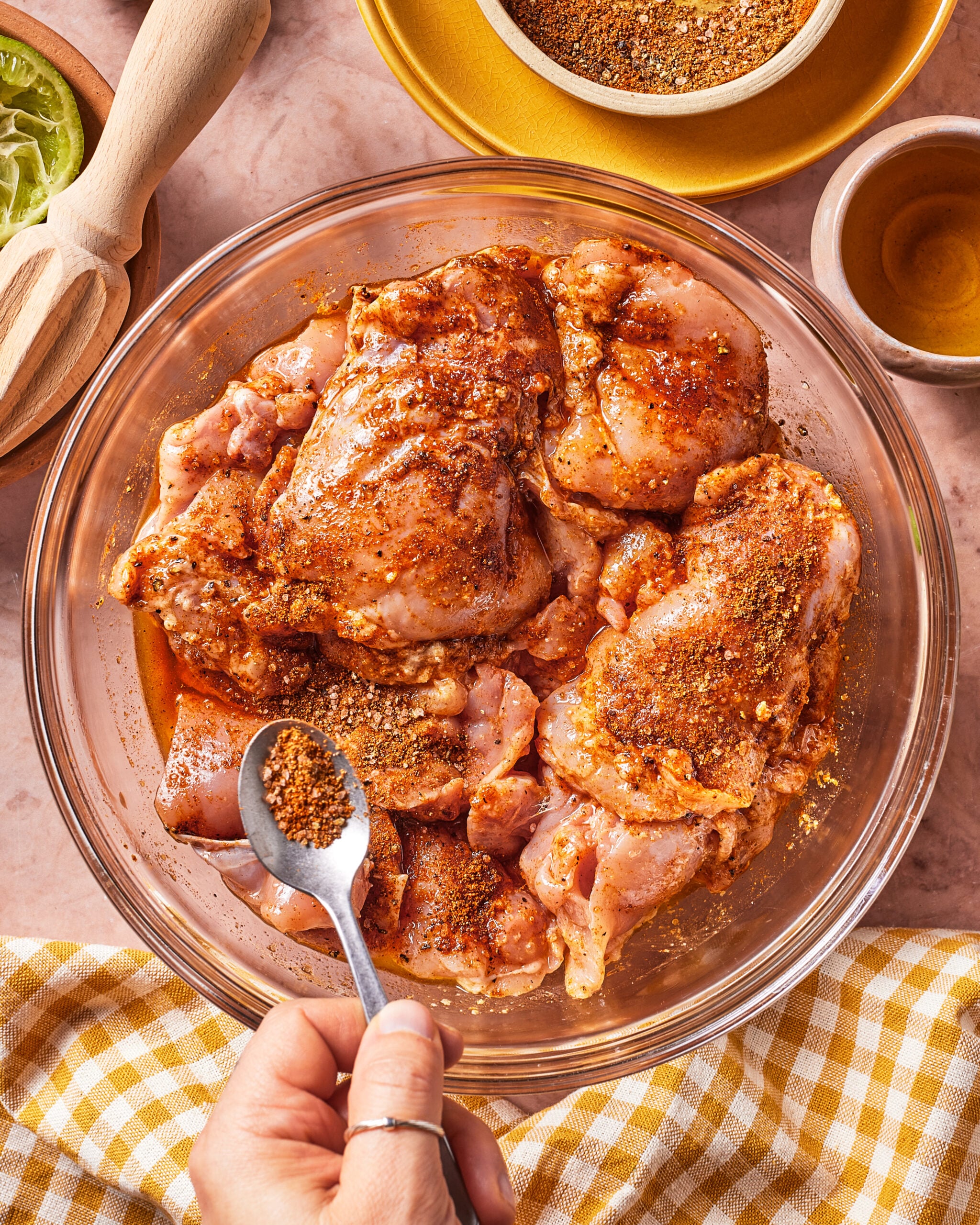 A hand sprinkles seasoning onto raw, marinated chicken pieces in a glass bowl on a kitchen counter with a yellow checkered cloth, spices, and a citrus reamer nearby.